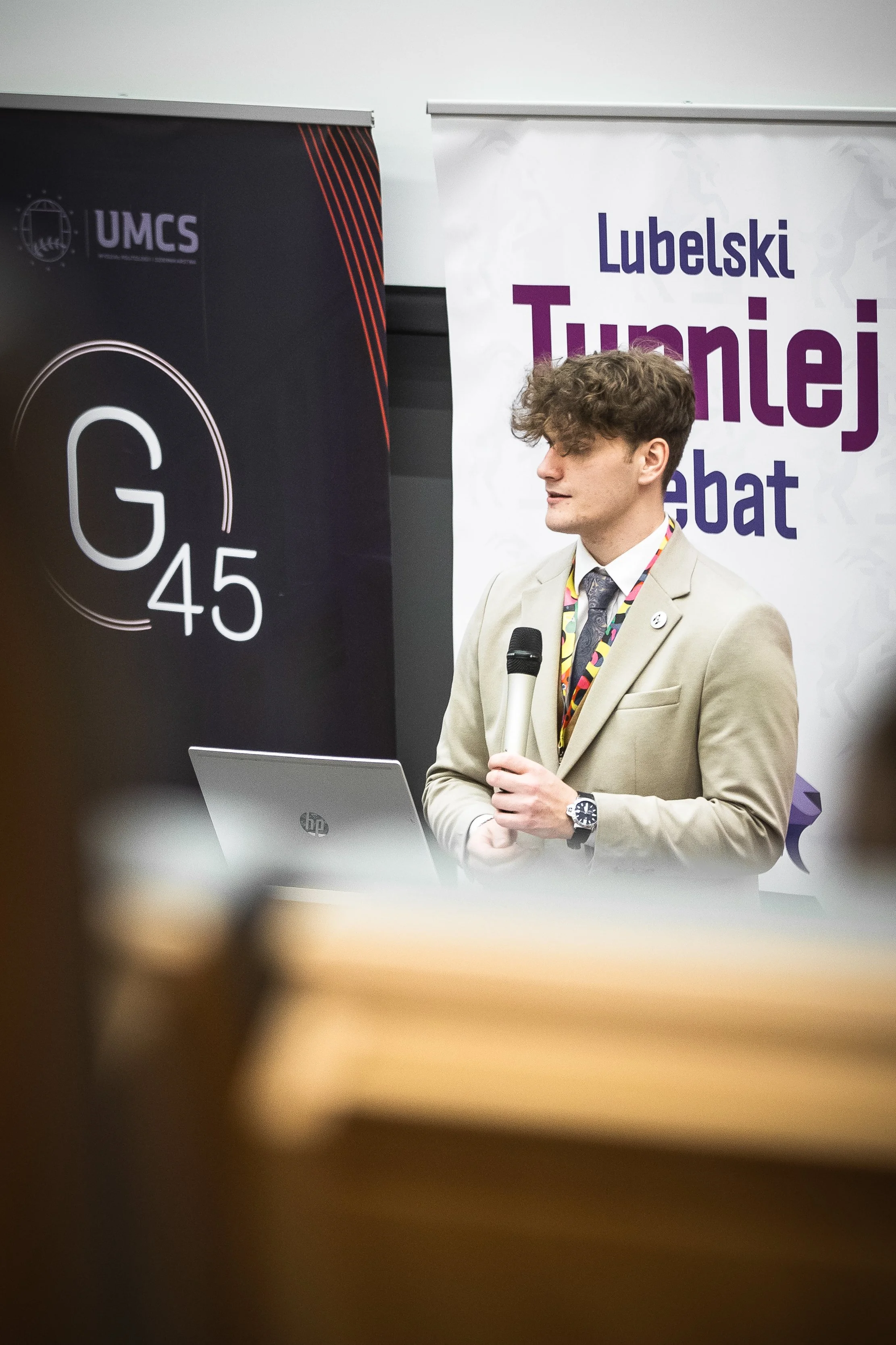 A young man in a beige suit holding a microphone, standing in front of two banners. One banner displays the logo for UMCS, and the other reads 'Lubelski Turniej Debat' in bold purple and blue letters.