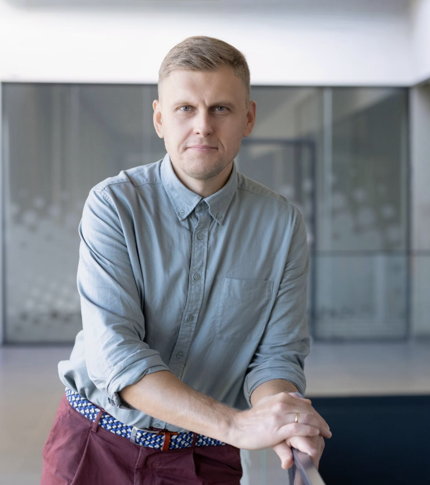 A man with short blond hair and blue eyes, wearing a light blue button-up shirt with rolled-up sleeves, burgundy pants, and a blue and white braided belt, standing indoors against a blurred background with modern decor.