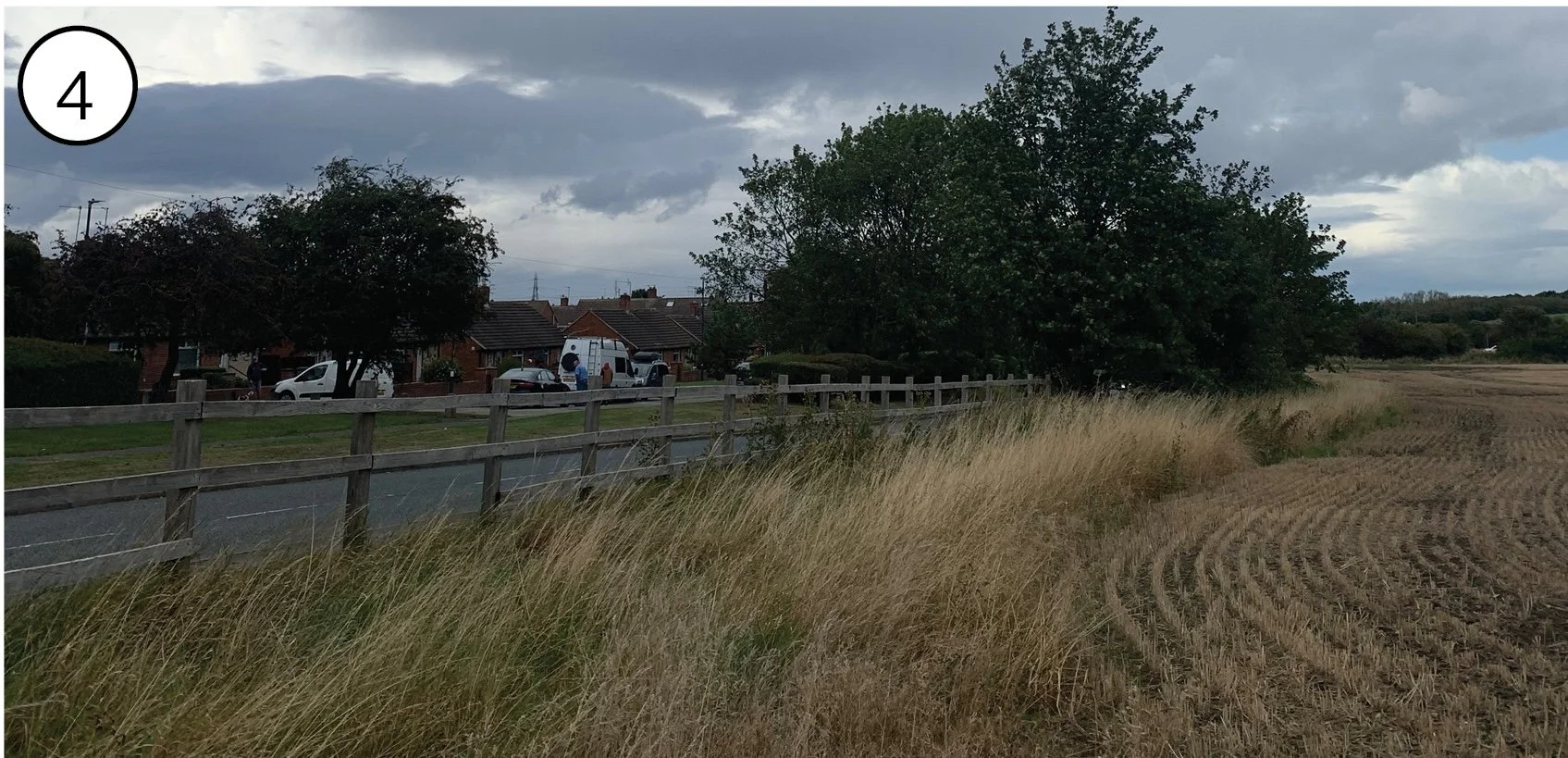 Rural scene with a plowed field, tall grass, a wooden fence, and a row of houses with cars parked outside, under a cloudy sky.