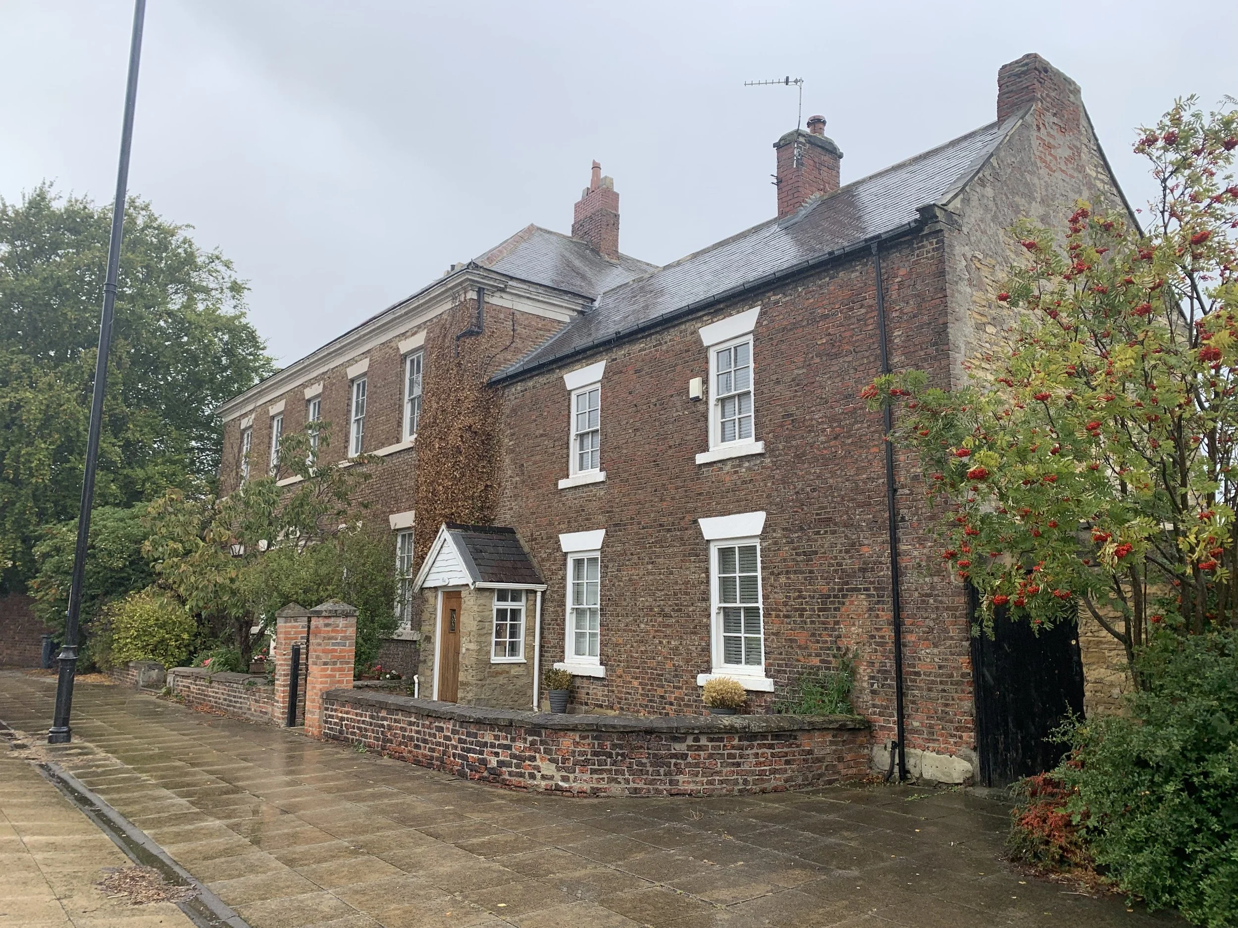 A traditional brick townhouse with white window frames, a small porch with a wooden door, a brick wall, and a garden with a tree bearing red berries. The street has a paved sidewalk and a lamppost.