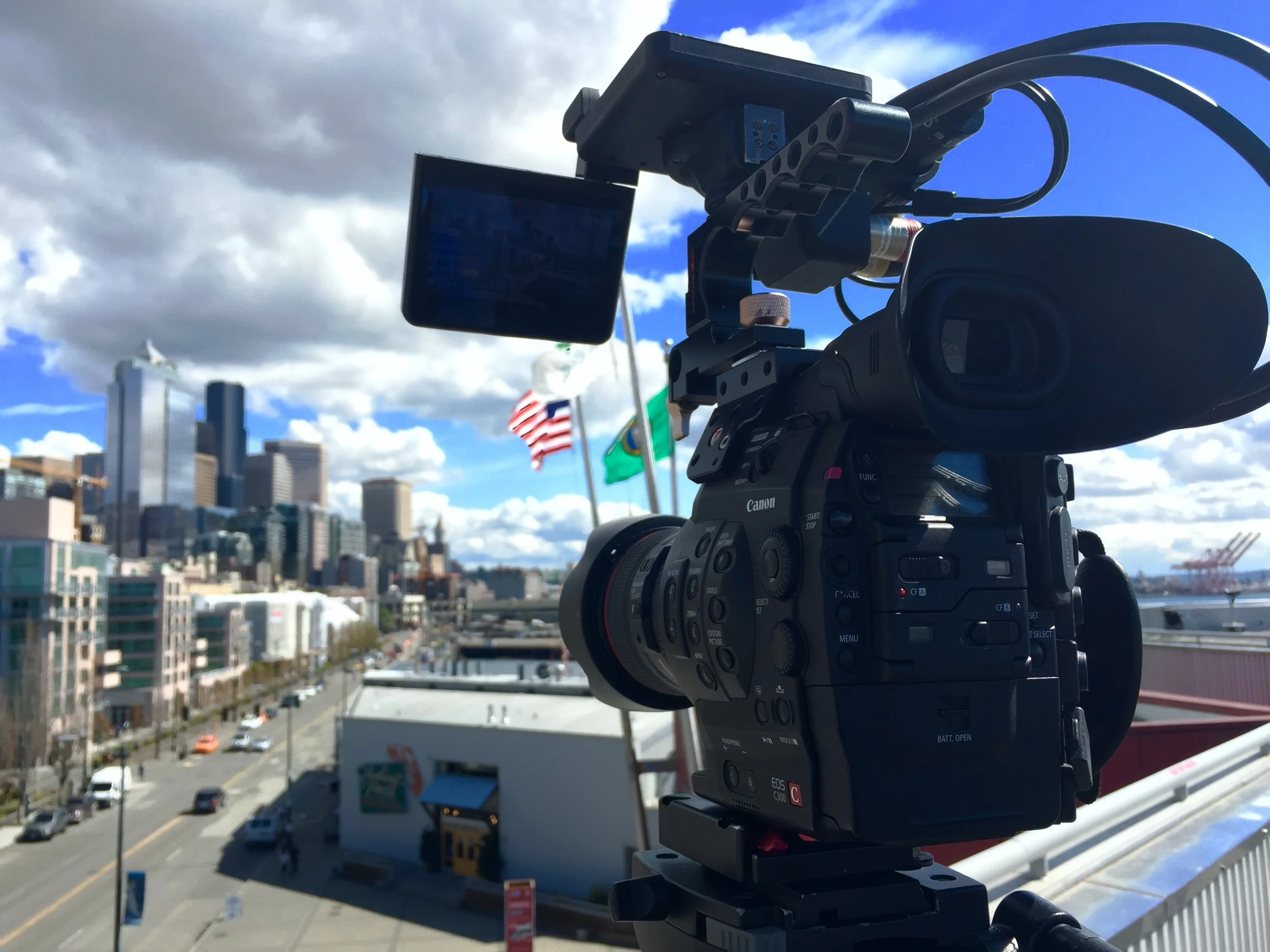 A professional video camera is positioned on a balcony or rooftop, capturing a cityscape with tall buildings, cars on the street, and flags fluttering in the wind against a partly cloudy sky.