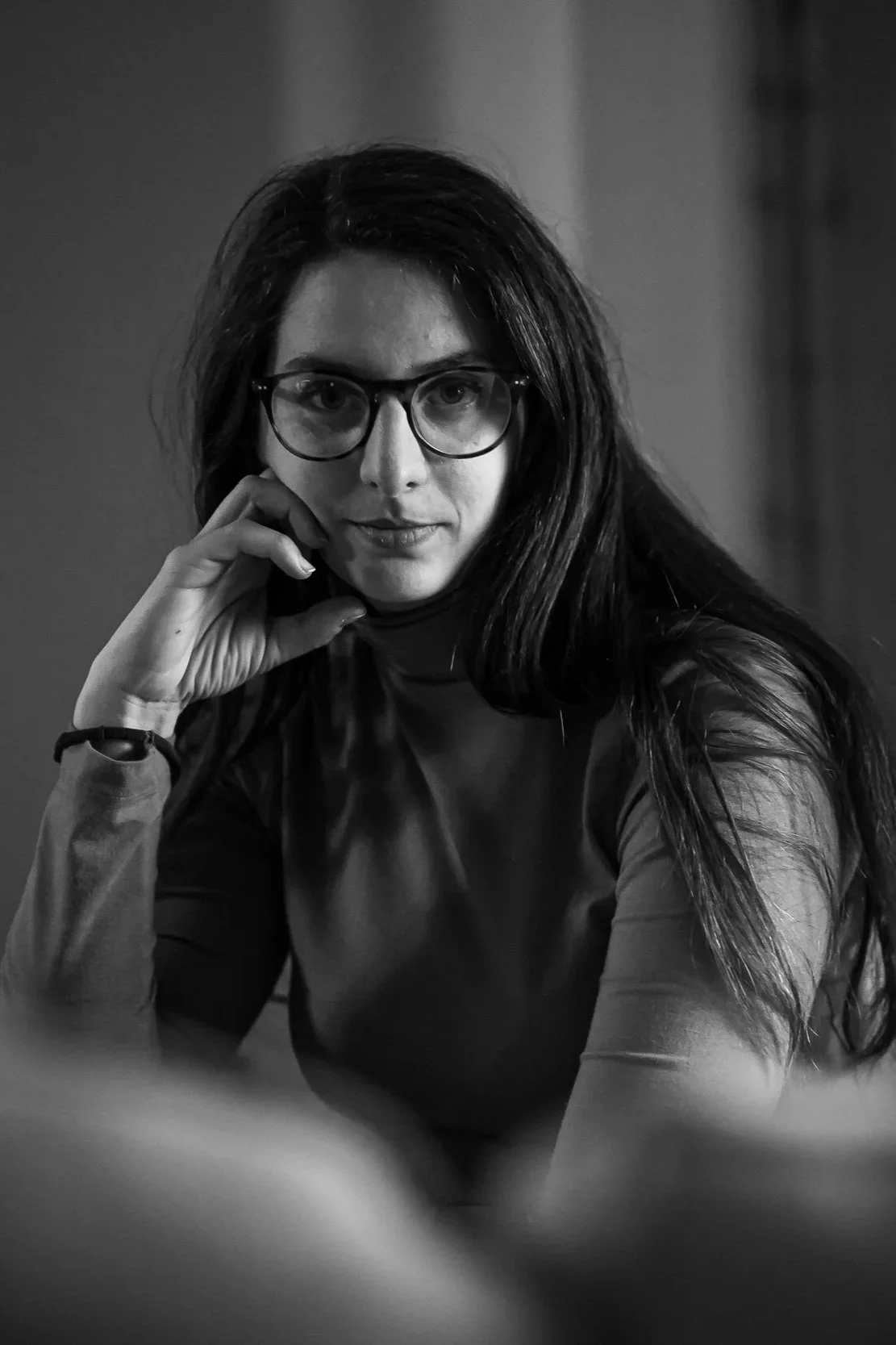 Black and white portrait of a woman with long dark hair and glasses, resting her chin on her hand, looking at the camera.