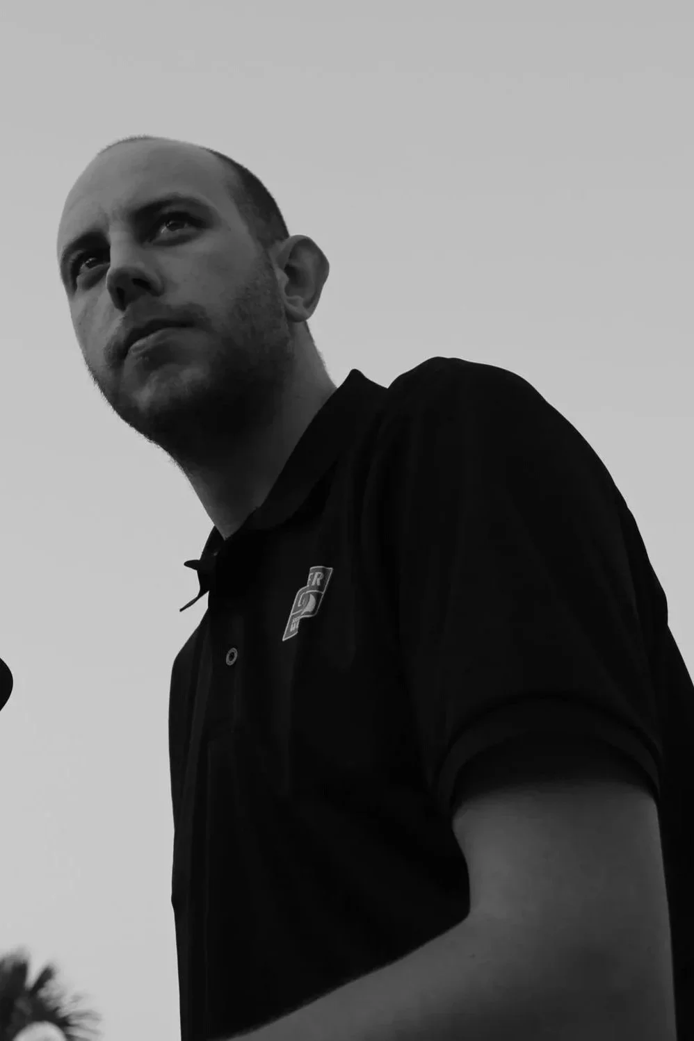 Black and white photo of a man with a shaved head and beard, wearing a dark collared shirt with a logo, looking off into the distance against a clear sky.