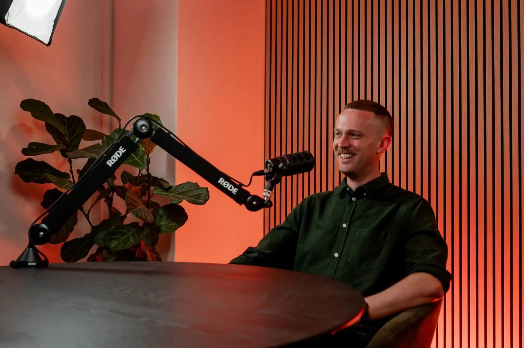 A man with short hair and a dark green button-up shirt smiling while sitting at a round table in a recording studio. A microphone labeled RØDE is positioned in front of him. There is a plant with large dark green leaves and a background of vertical wooden slats on the wall.
