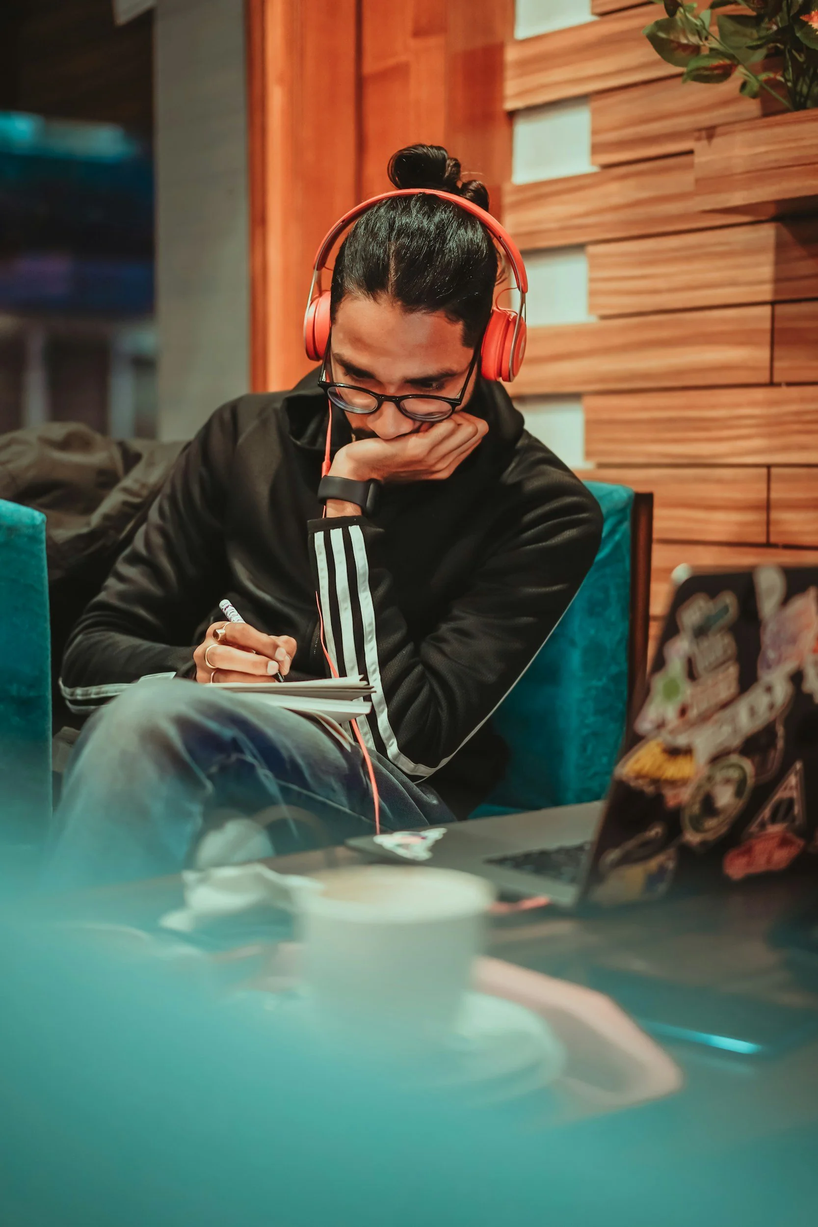 A young man with glasses and dark hair tied in a bun, wearing headphones and a black jacket, is sitting down, looking down at a notebook and holding a pen. He appears to be in a cozy indoor space with wooden wall paneling and a plant in the background.