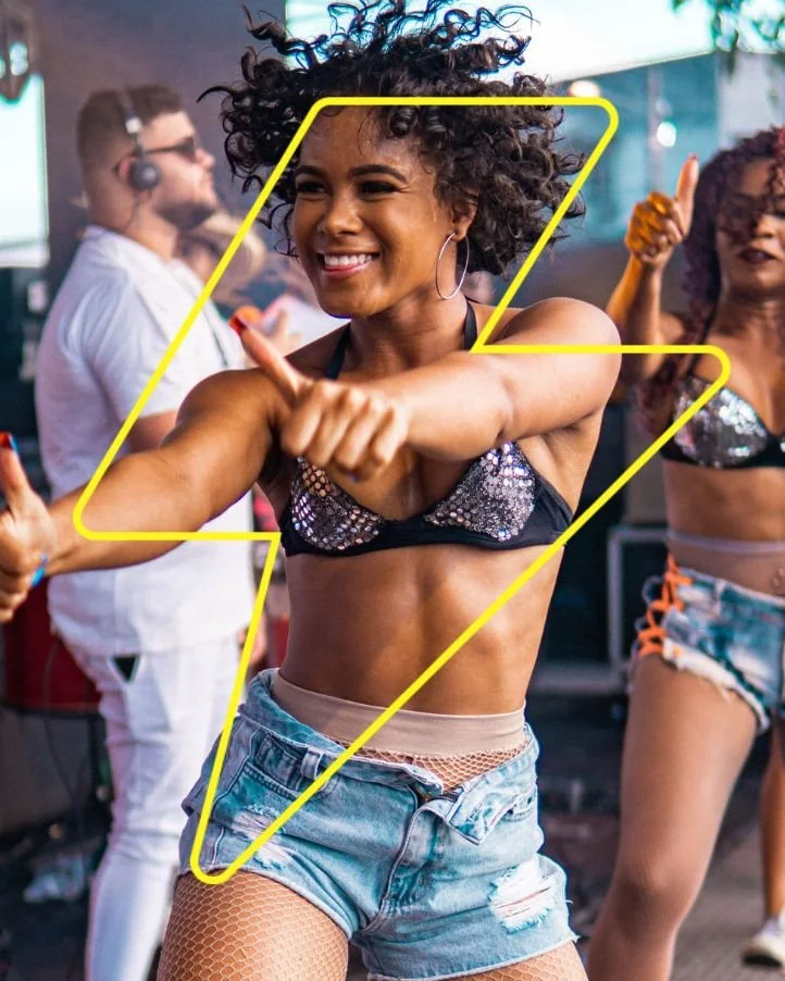 A woman dancing at a lively party or club with people in the background, wearing a sparkly top, denim shorts, fishnet stockings, and large hoop earrings, smiling and giving a thumbs-up.