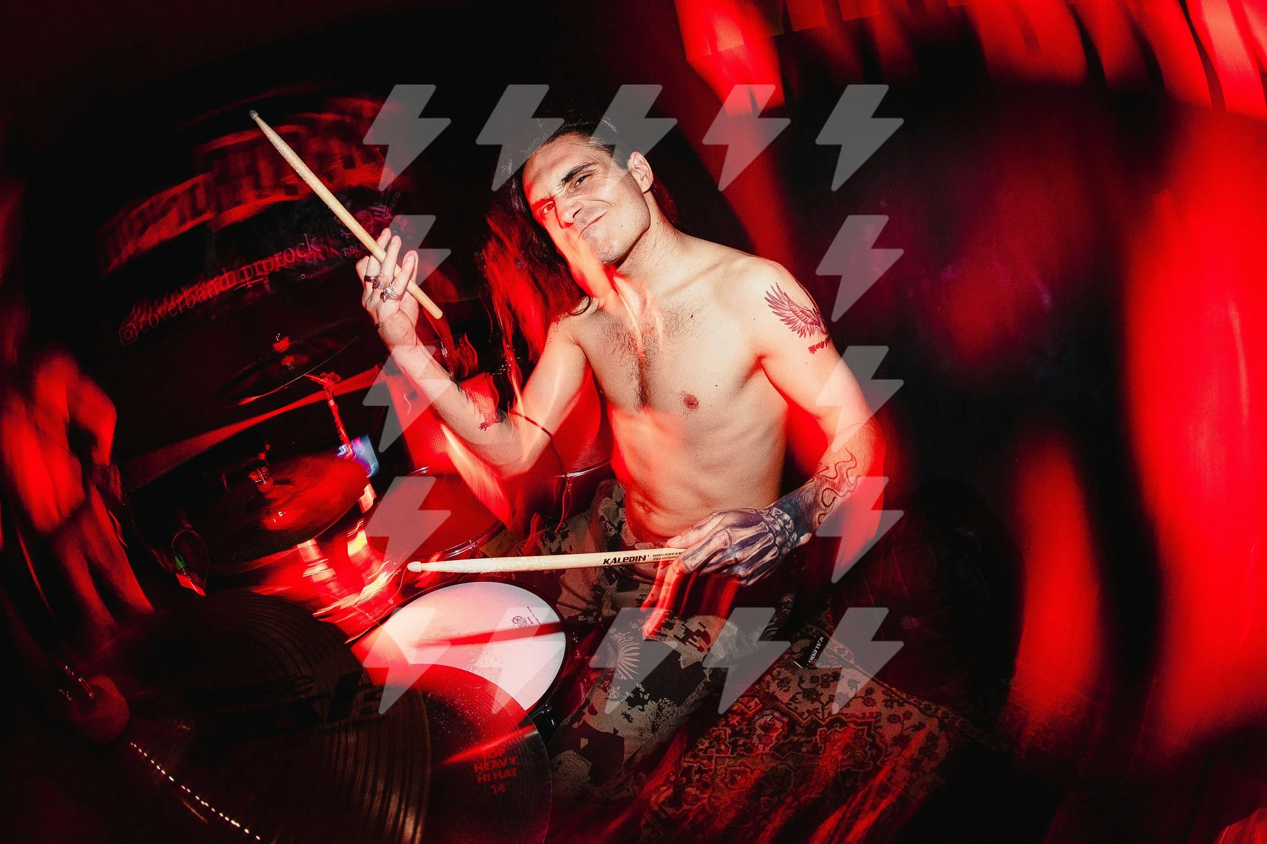 A shirtless man with long hair and tattoos, holding drumsticks, smiling in a dimly lit red room with lightning bolt graphics overlay.