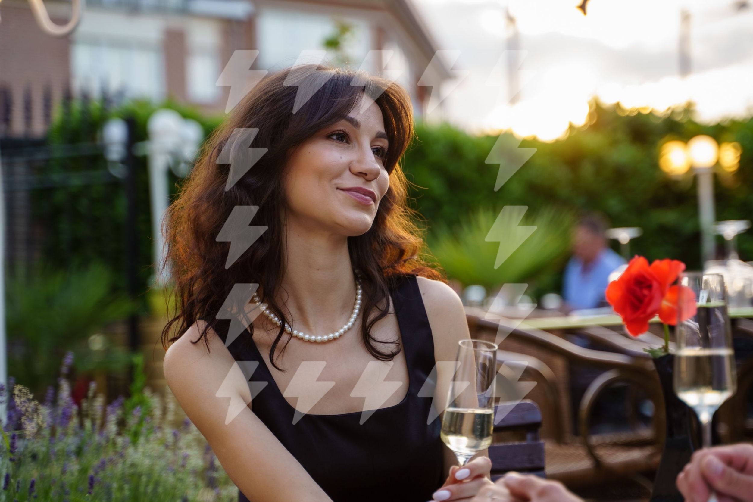 A woman with dark wavy hair wearing a black dress and pearl necklace, smiling and holding a glass of champagne at an outdoor event during sunset.