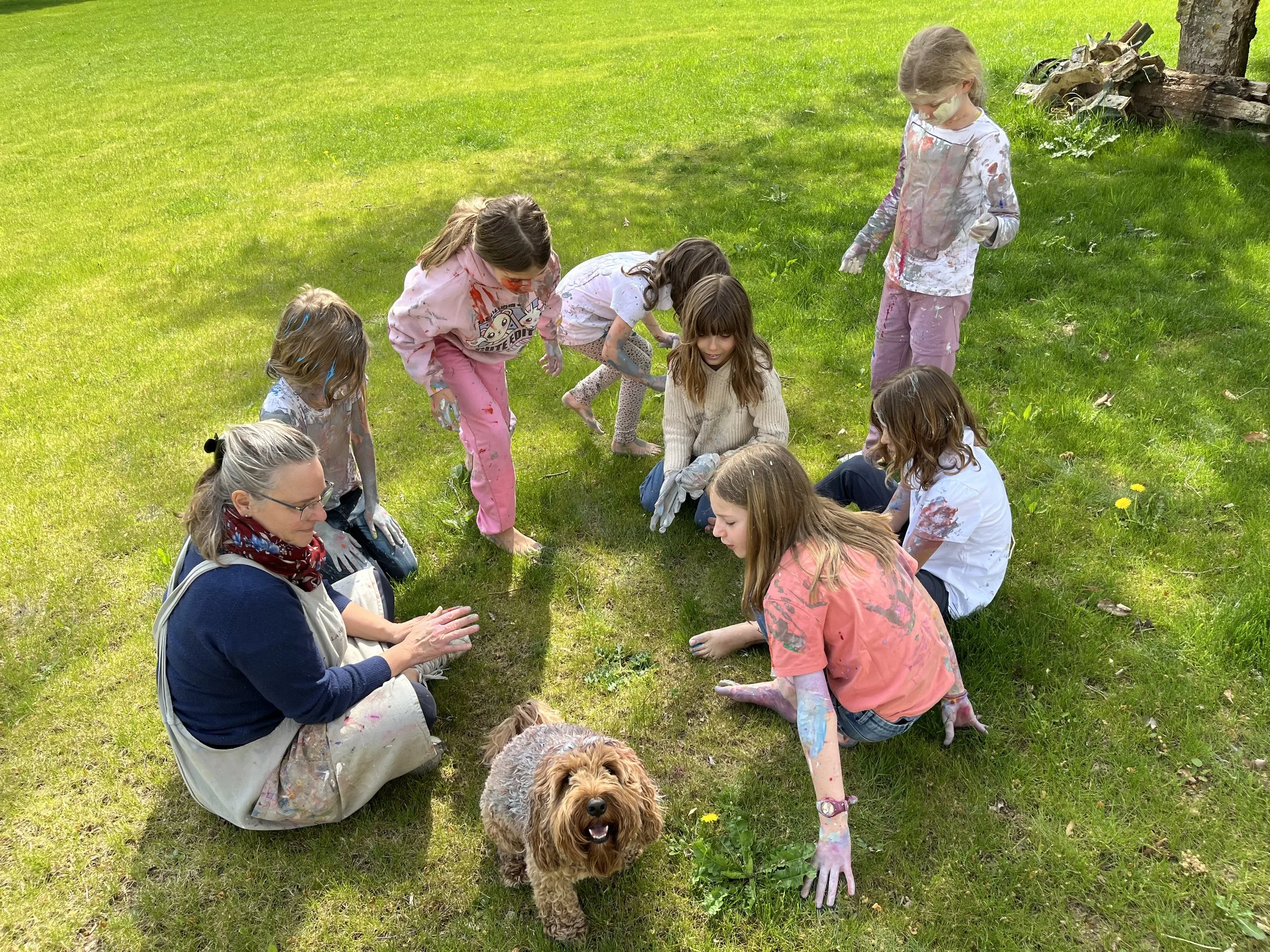 Group of children and an adult sitting and kneeling on grass outdoors, covered in colorful paint, with a brown dog in the foreground.