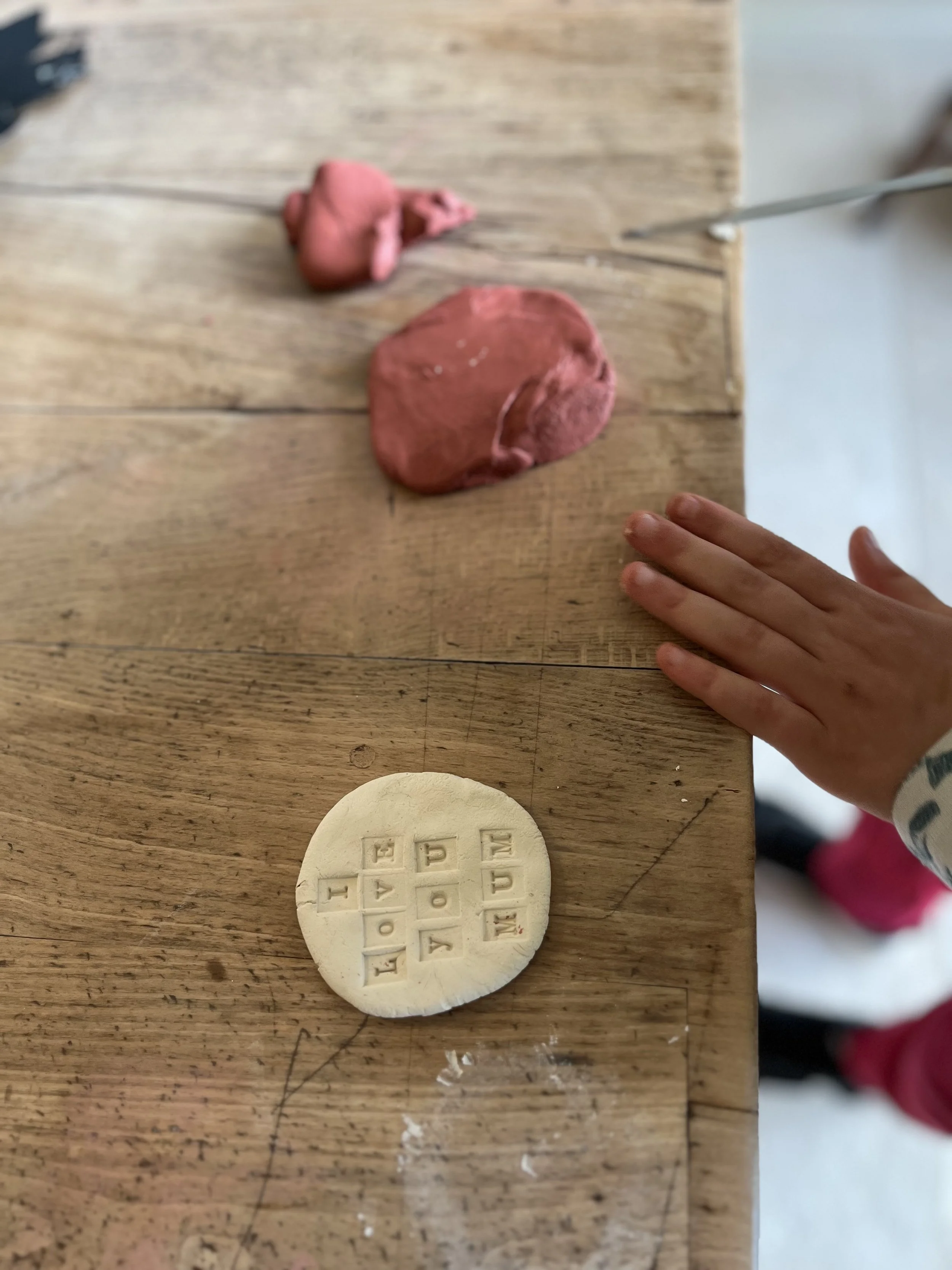A wooden table with pink playdough, a white round cookie with embossed letters and numbers, and a child's hand with fingers resting on the table.
