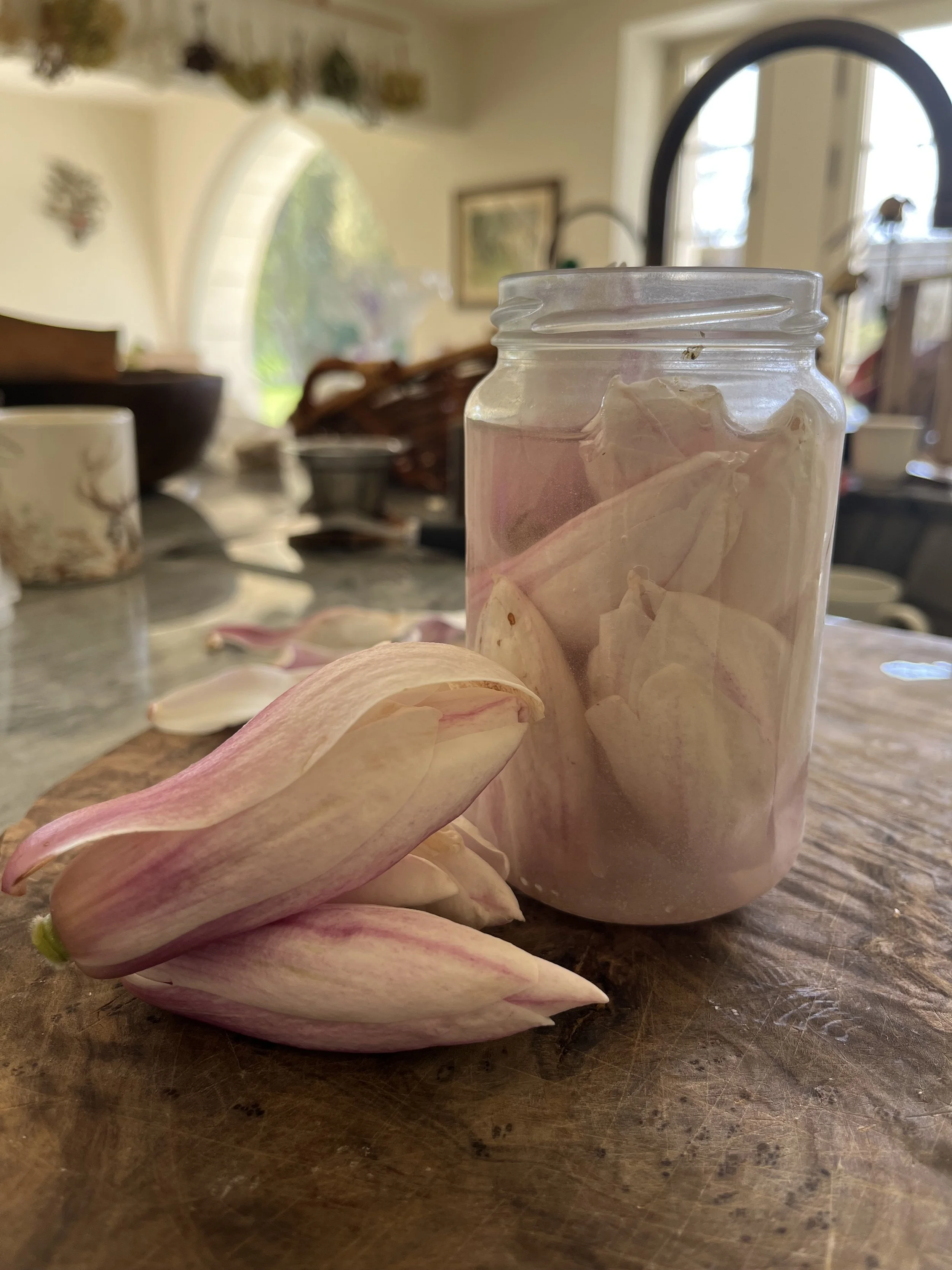 A glass jar filled with dried flower petals, placed on a wooden table. There are more petals scattered on the table around the jar.