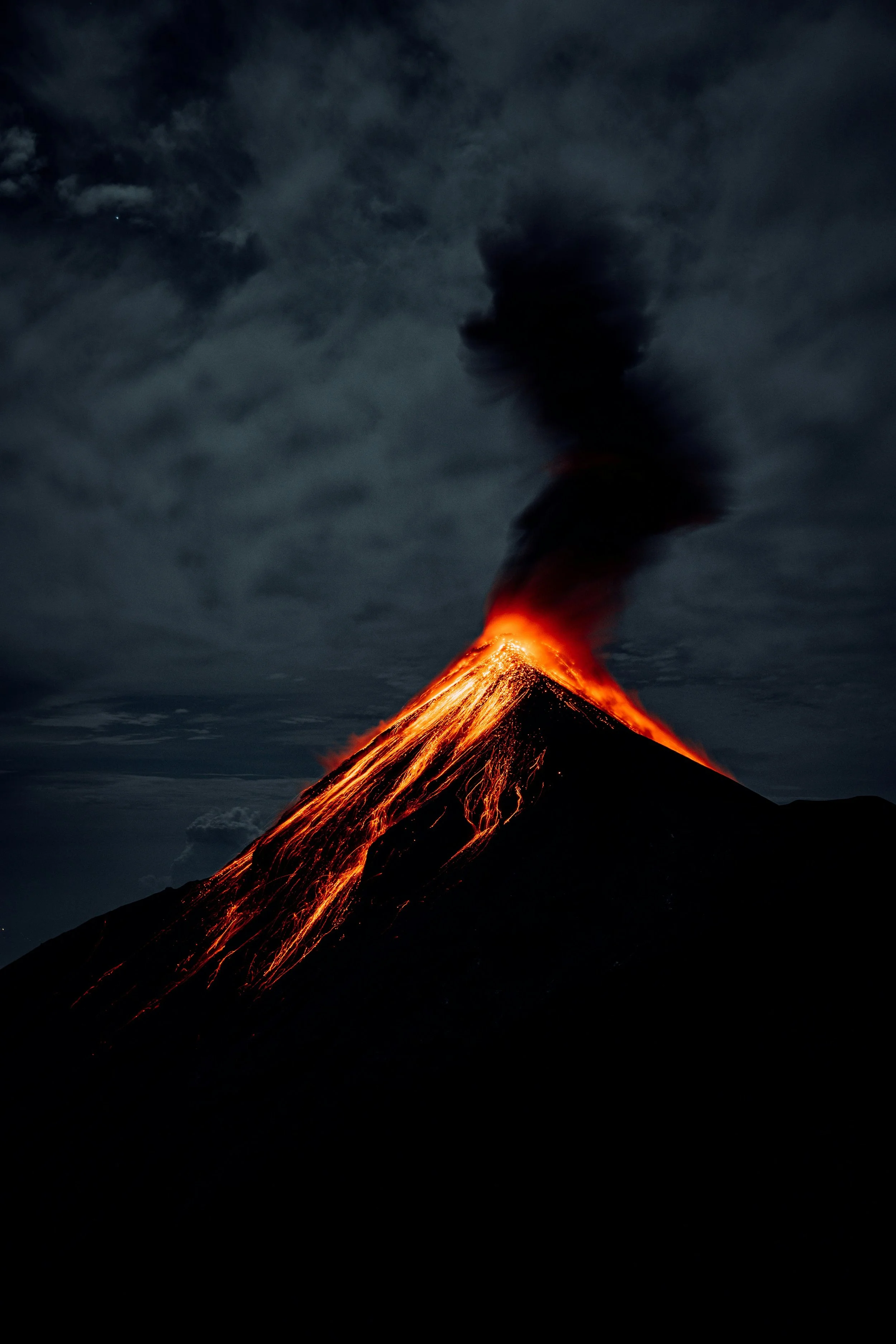A volcanic eruption at night with bright lava flowing down the sides and dark smoke billowing into the cloudy sky.