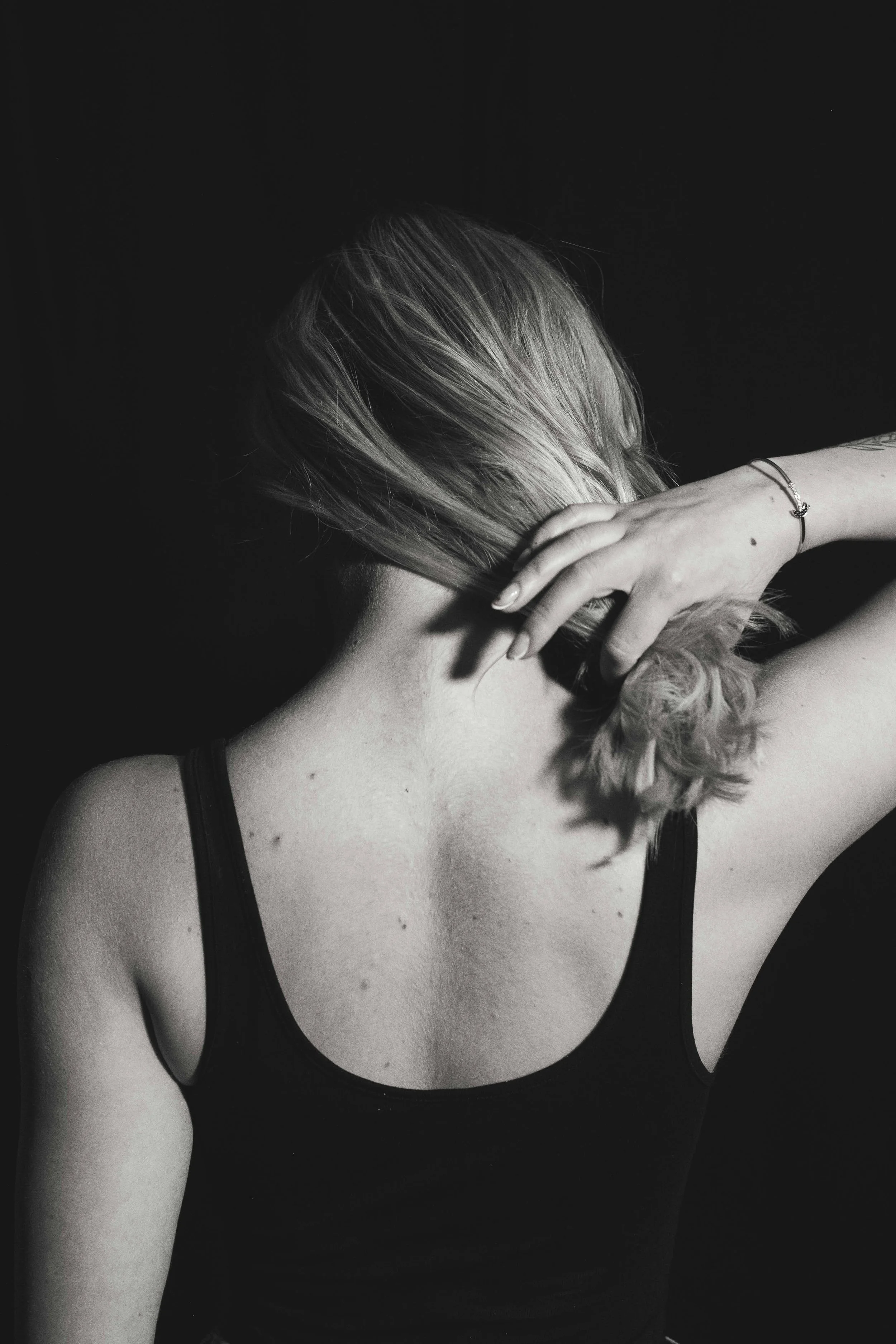Black and white photo of a woman with wavy hair, wearing a spaghetti strap top, with her hand resting on her neck and face, against a dark background.