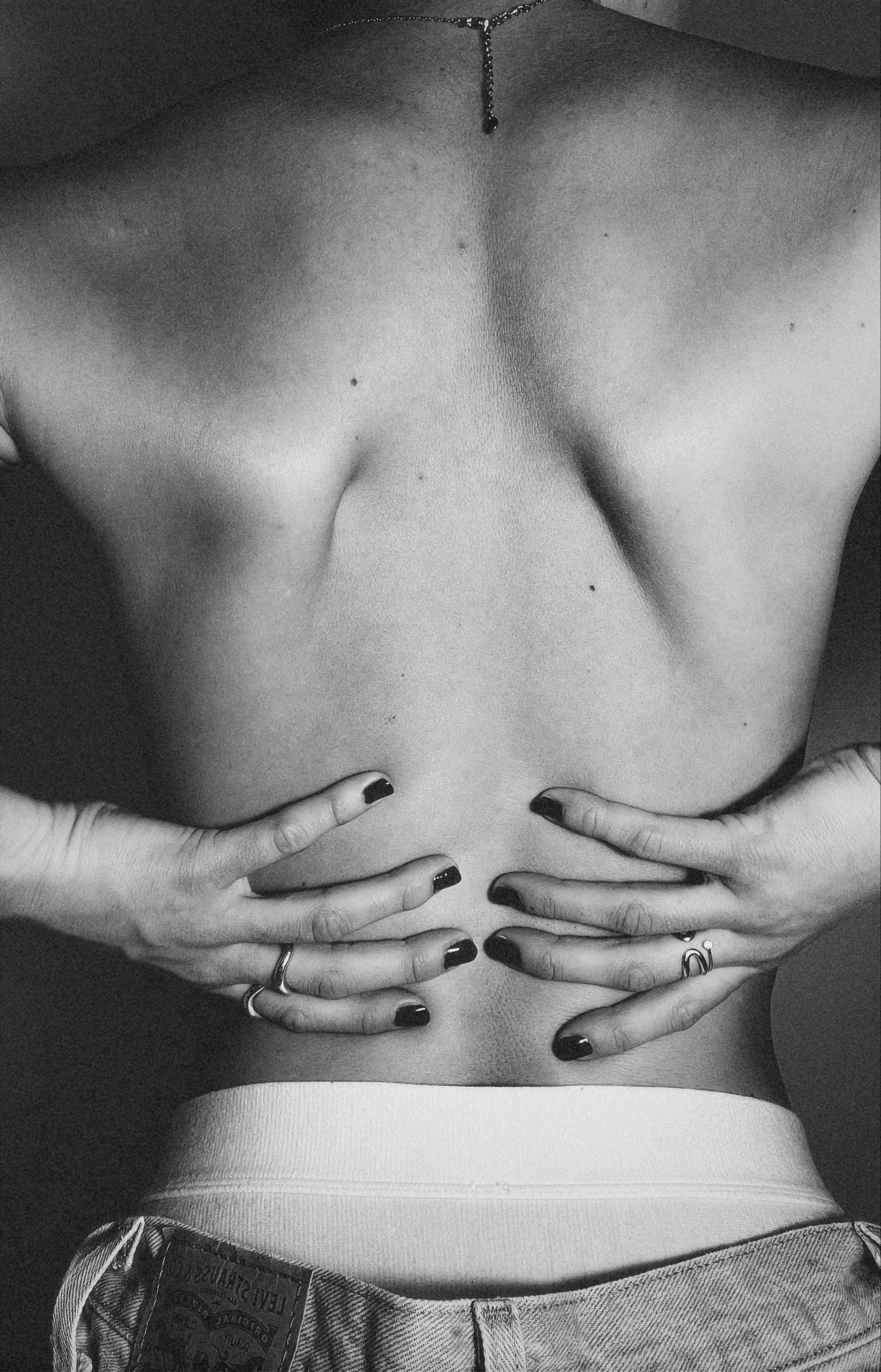 Black and white photo of a person's bare chest with hands placed on their stomach. The person is wearing jeans and a necklace.