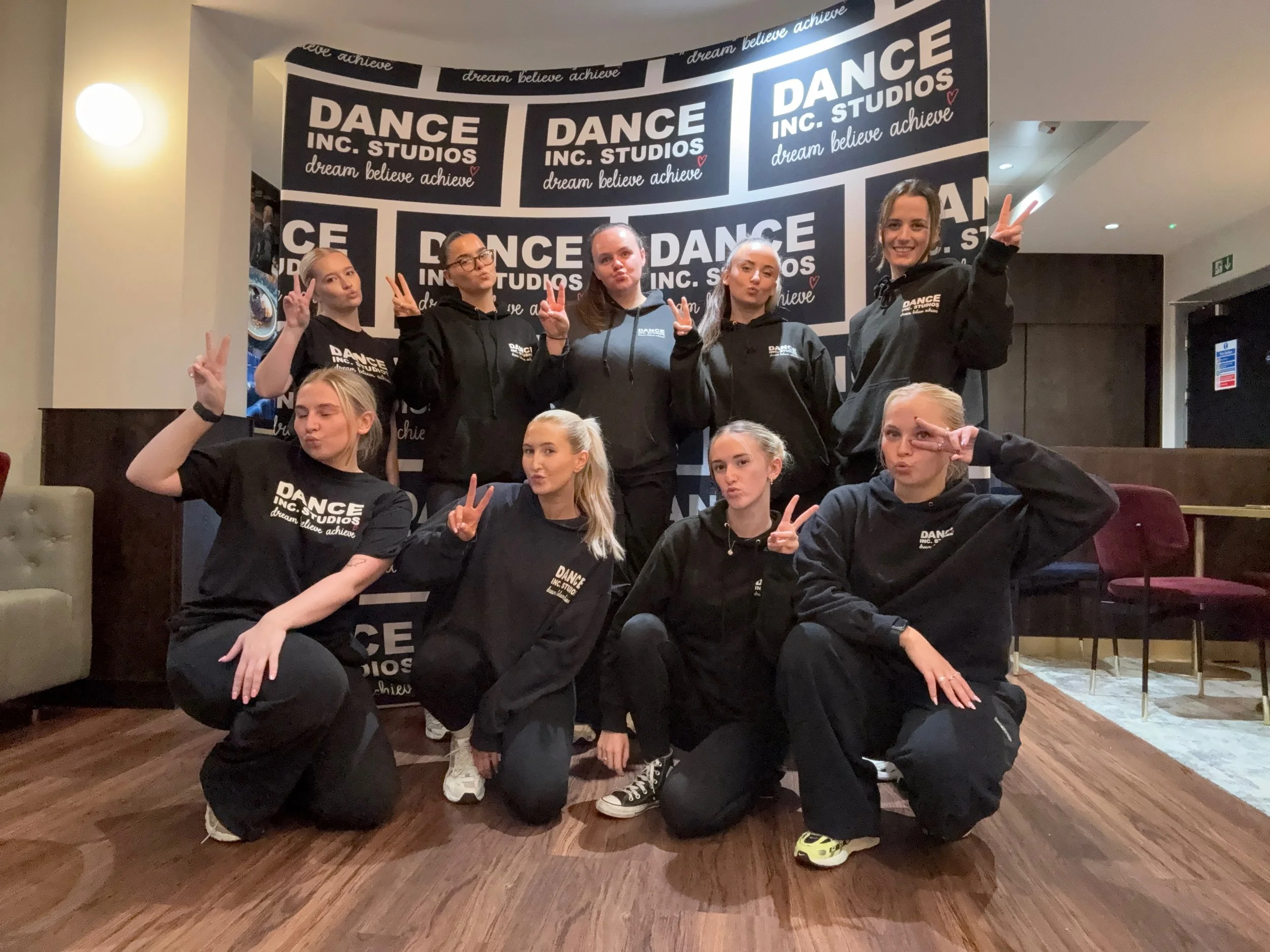 Group of young women in black hoodies with 'Dance Inc Studio' logo, posing with peace signs and making silly faces in front of a black and white dance studio sign.