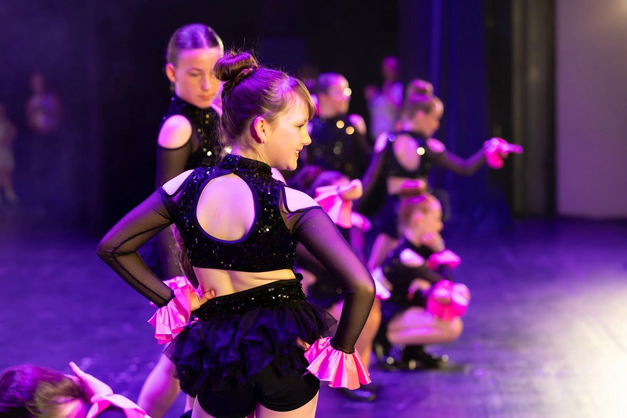 Young girls in black dance costumes with pink ruffles practicing on stage under purple lighting.
