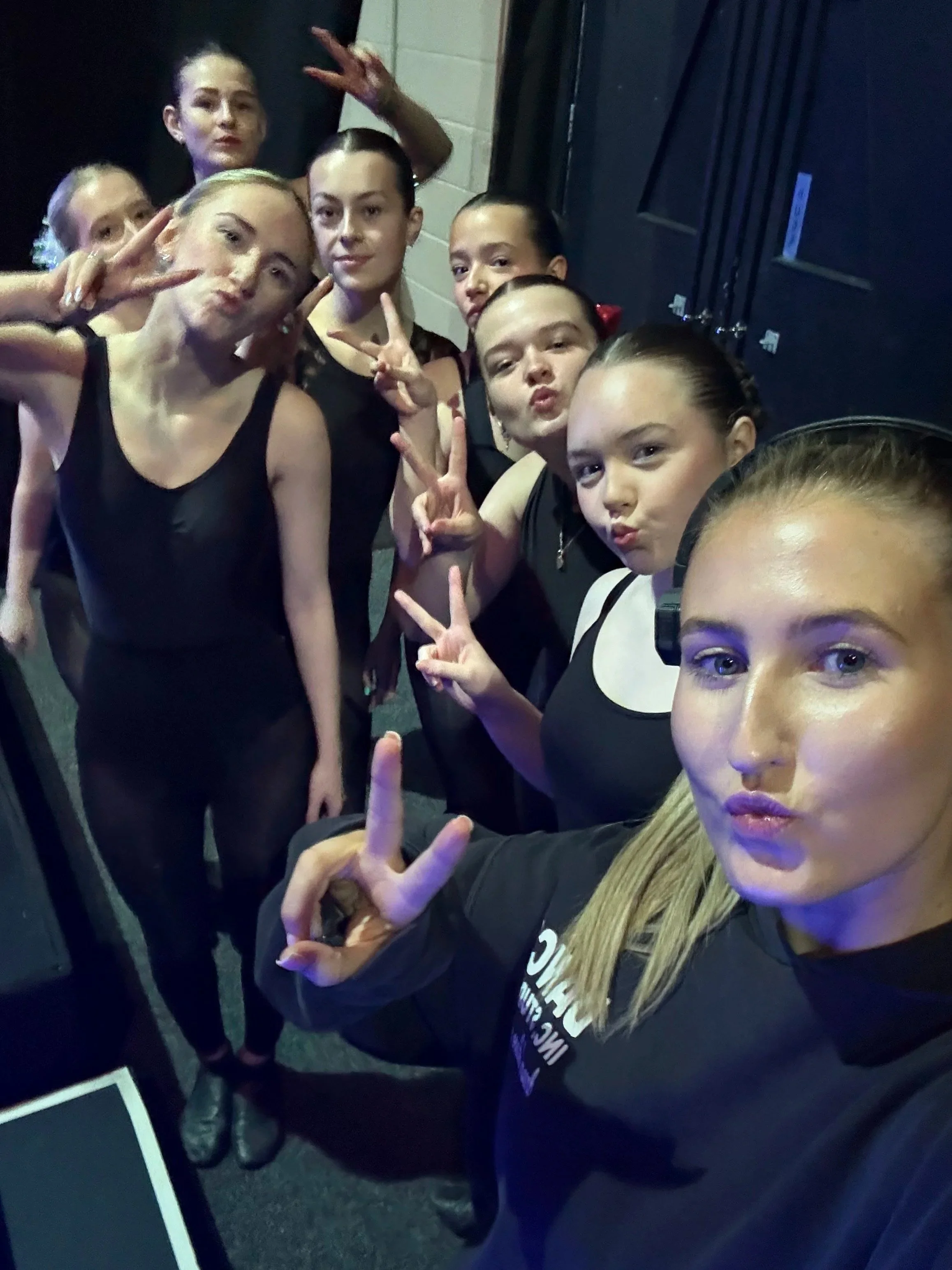 Group of young female dancers taking a selfie backstage, some making peace signs and pout faces, dressed in black dance costumes.