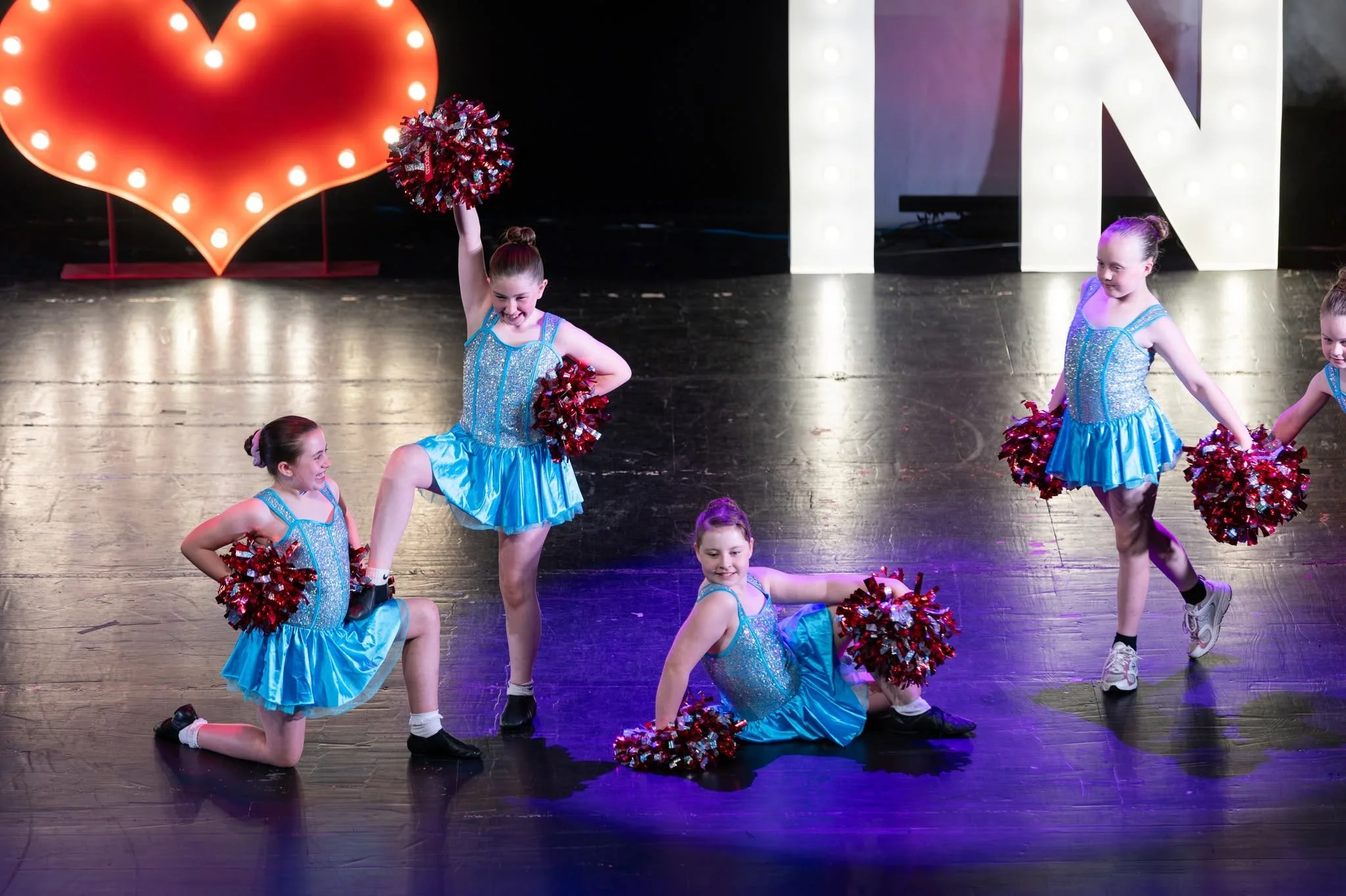 A group of young girls in silver and black tutus performing a ballet on stage, with some fixing their hair.