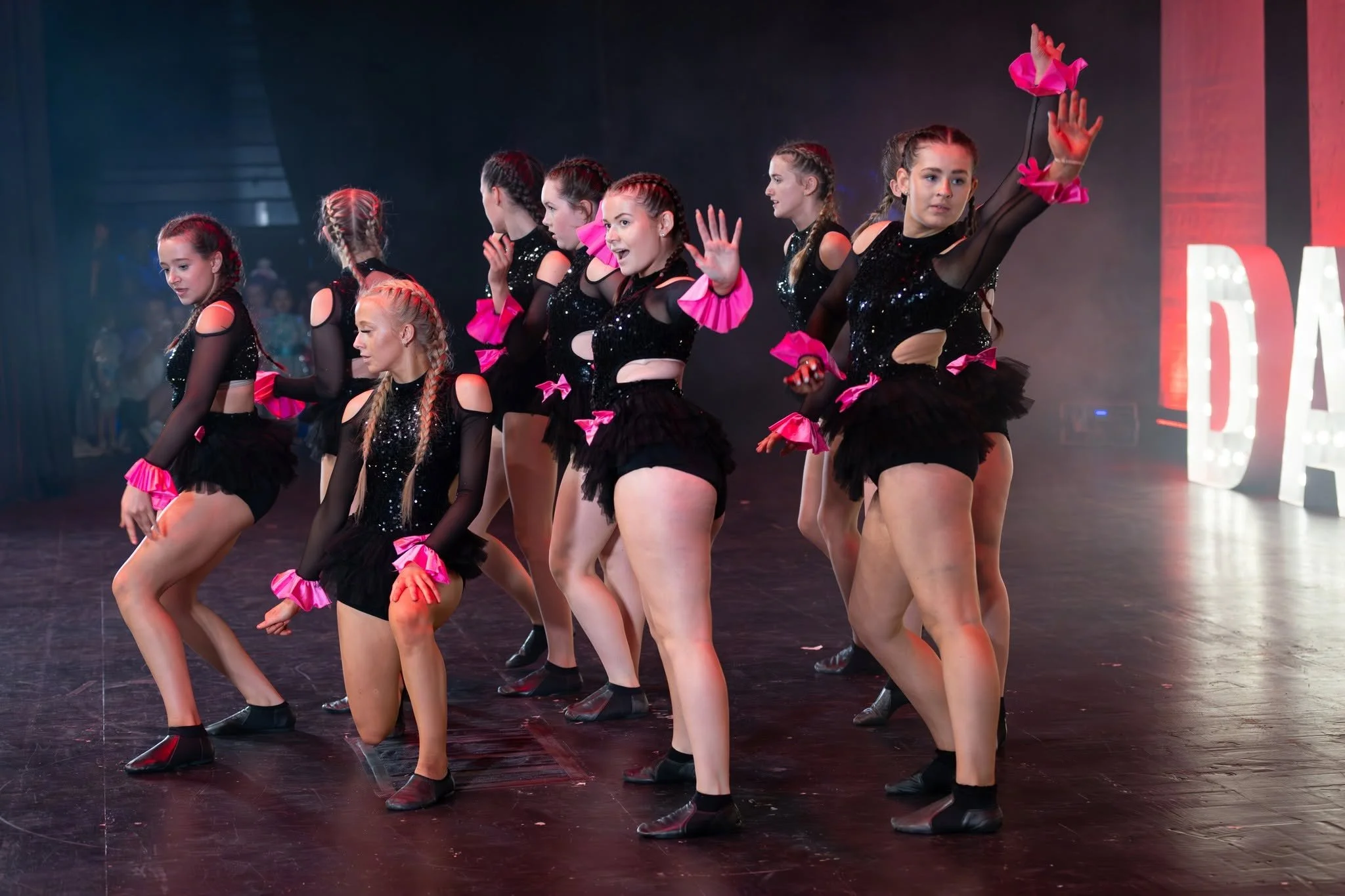 Group of young female dancers in black costumes with pink accents performing a dance routine on stage.