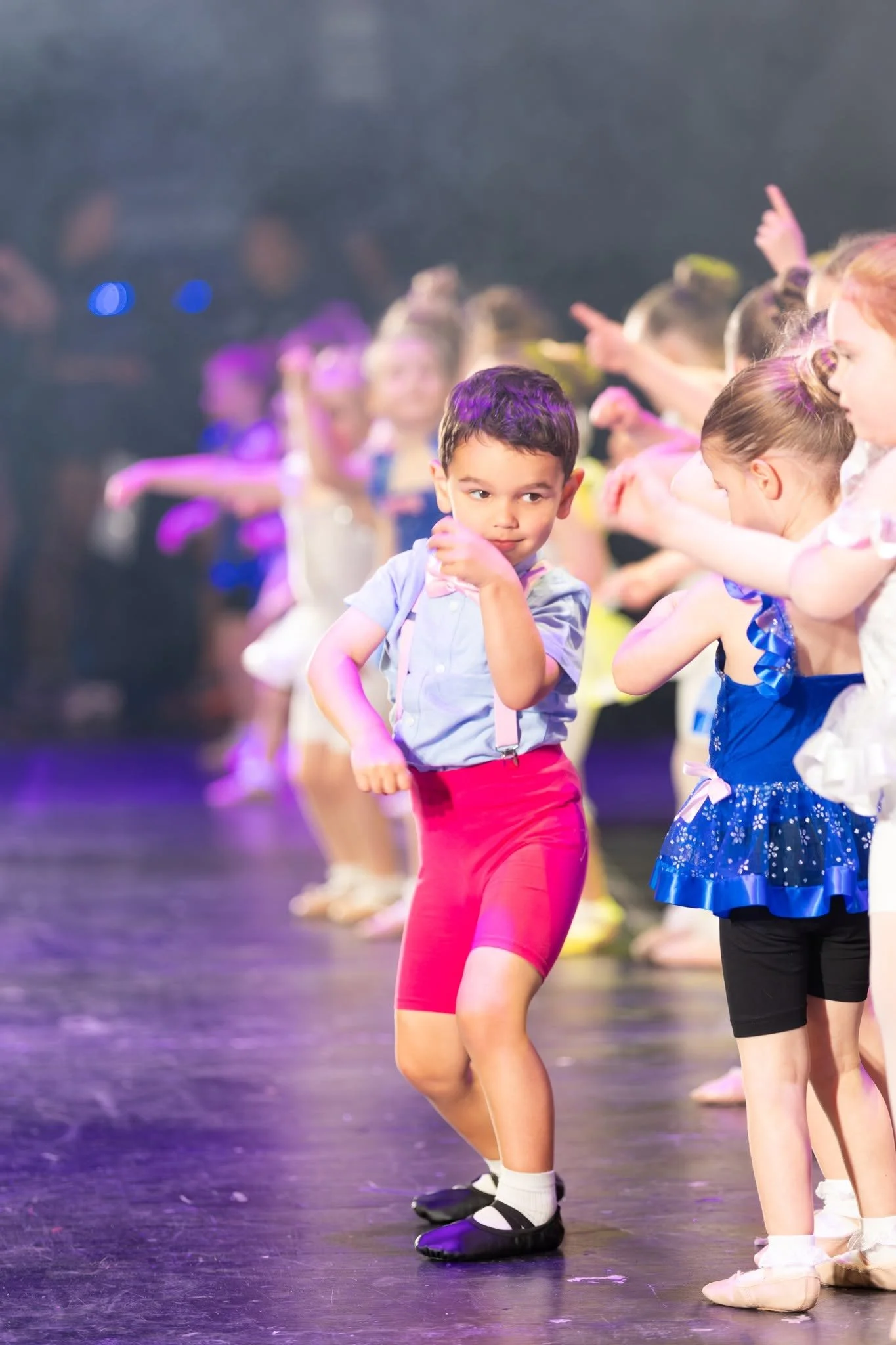 Young girl in a white ballet costume with silver embellishments and a tulle skirt, performing ballet on stage with other dancers in the background, under stage lights.