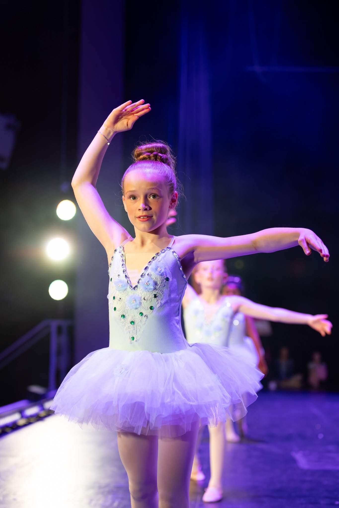 Young girl in a white ballet costume with silver embellishments and a tulle skirt, performing ballet on stage with other dancers in the background, under stage lights.
