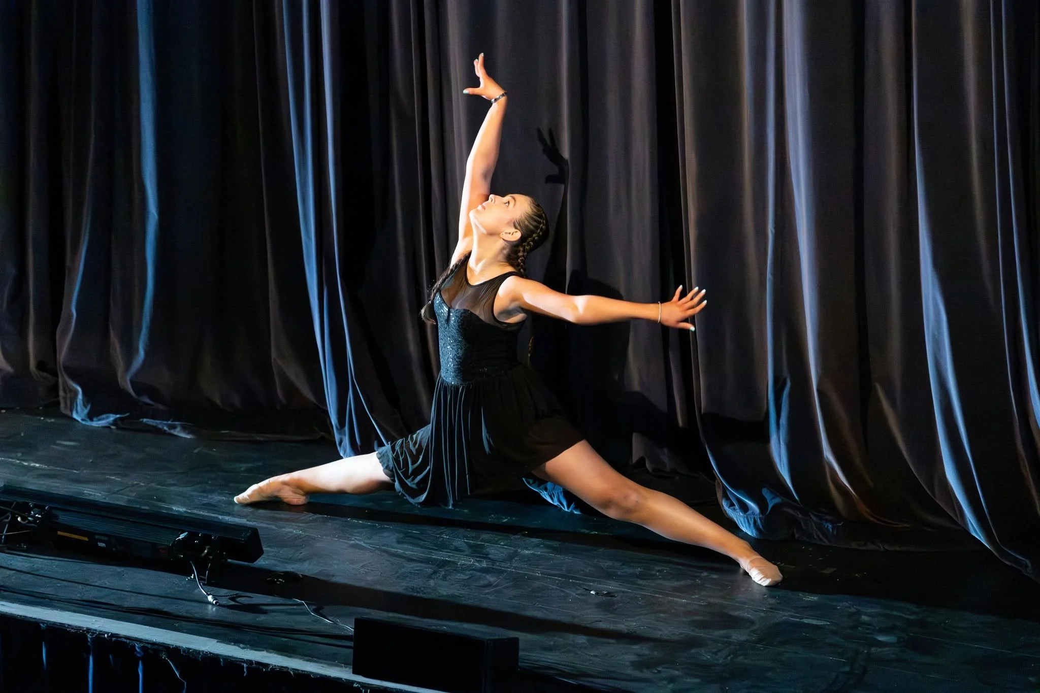 A young woman in a black dance costume is performing a dance move on stage, kneeling on one knee with her other leg extended behind her, with arms gracefully raised and head tilted back, against a background of dark curtains.