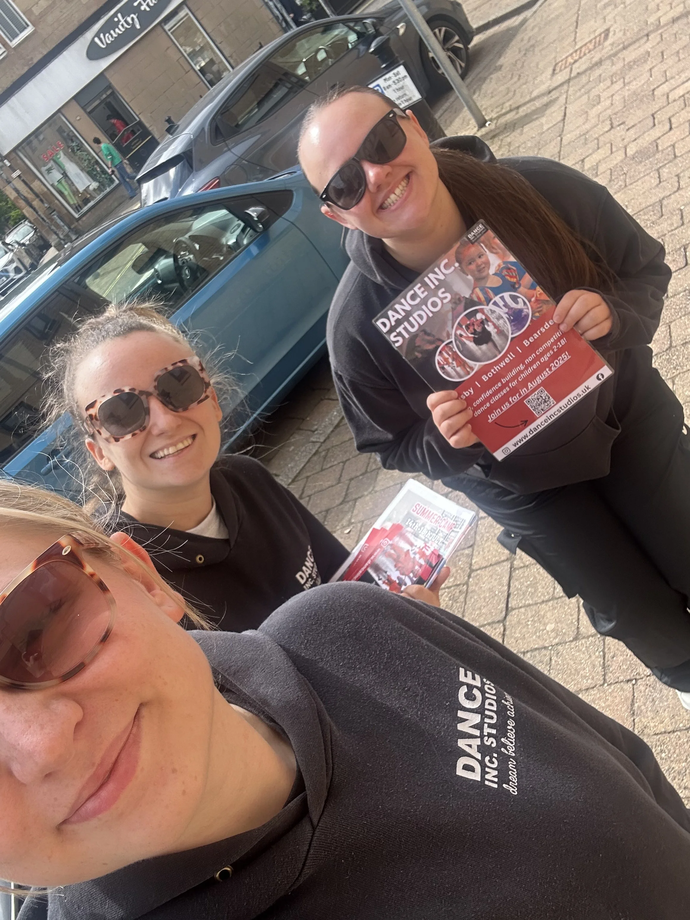 Smiling woman with sunglasses taking a selfie on a city sidewalk, holding a flyer for dance classes at Dance Inc. Studios, with parked cars and storefronts in the background.