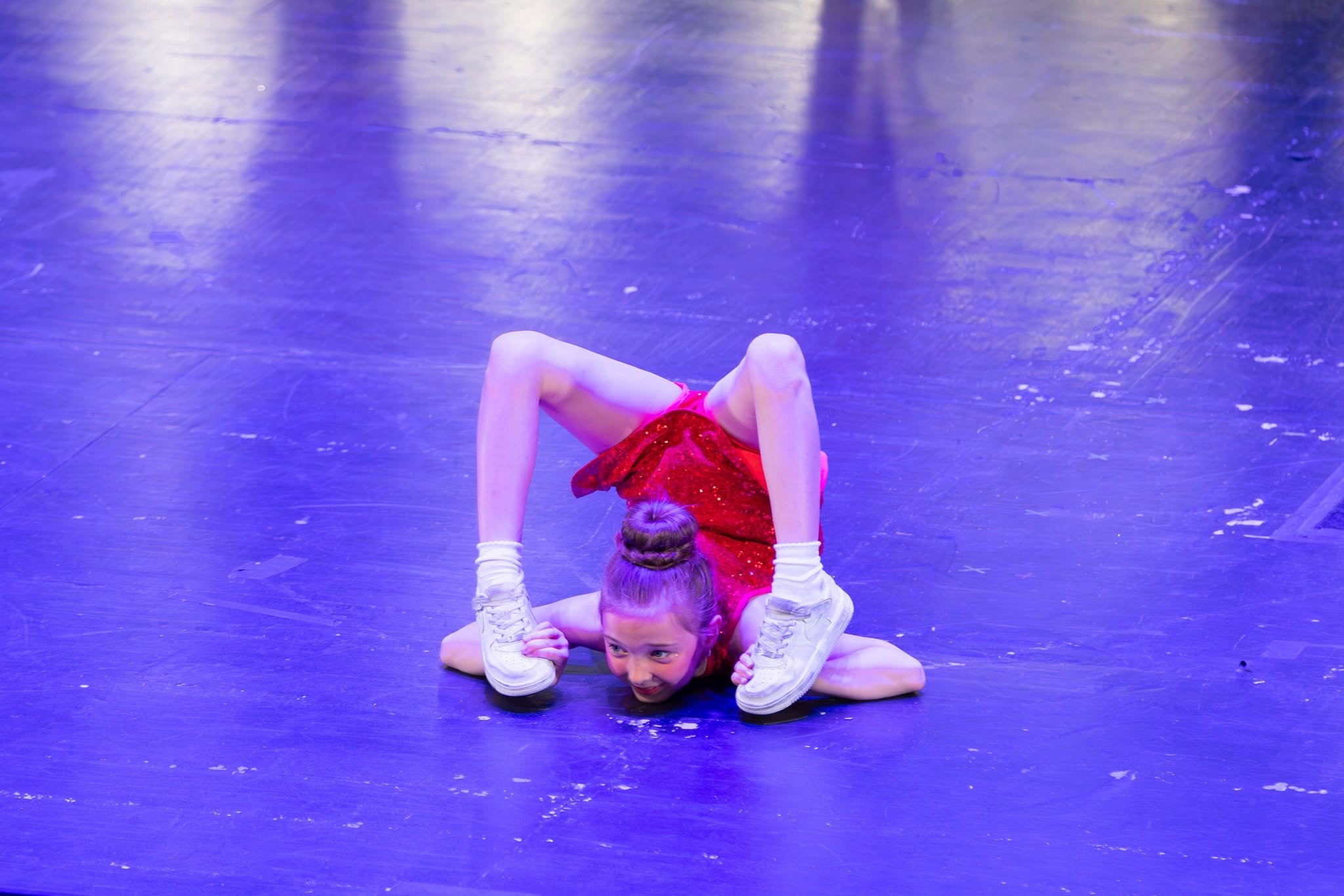 Two young female dancers performing an intricate pose on a stage with a shiny, purple-lit floor. One girl is lying on the floor with her head resting on her hands and smiling, while the other is balanced above her, supporting herself with her arms and legs bent.