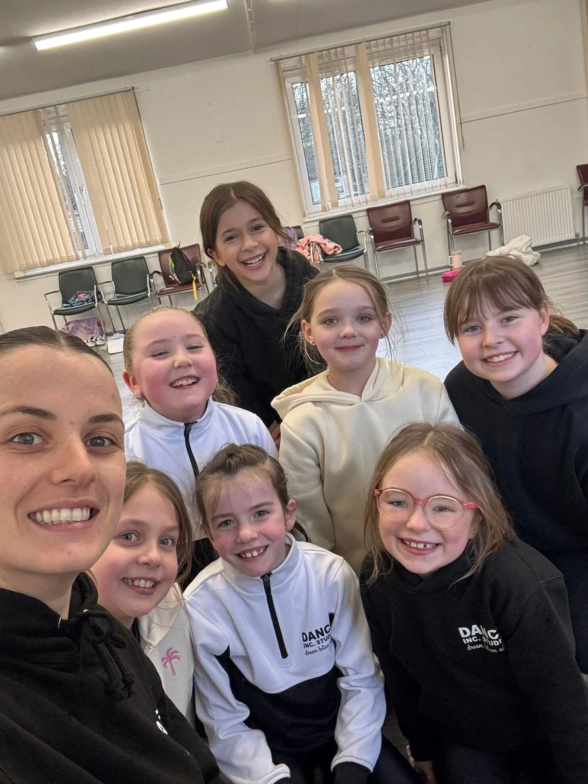 Group of smiling children and a young woman taking a selfie in a room with large windows and beige vertical blinds.