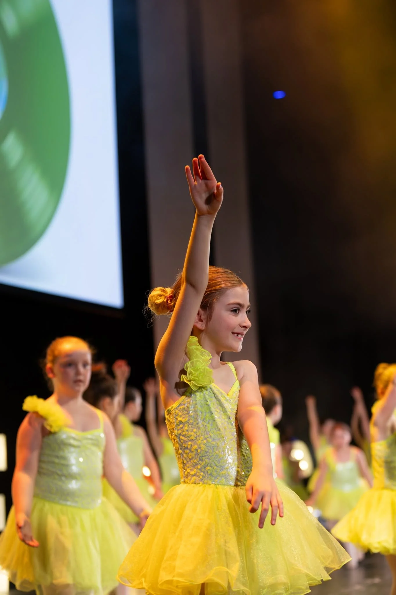 Young girl in a light-colored dress with a pink bow in her hair dancing on stage with others, raising her arms.