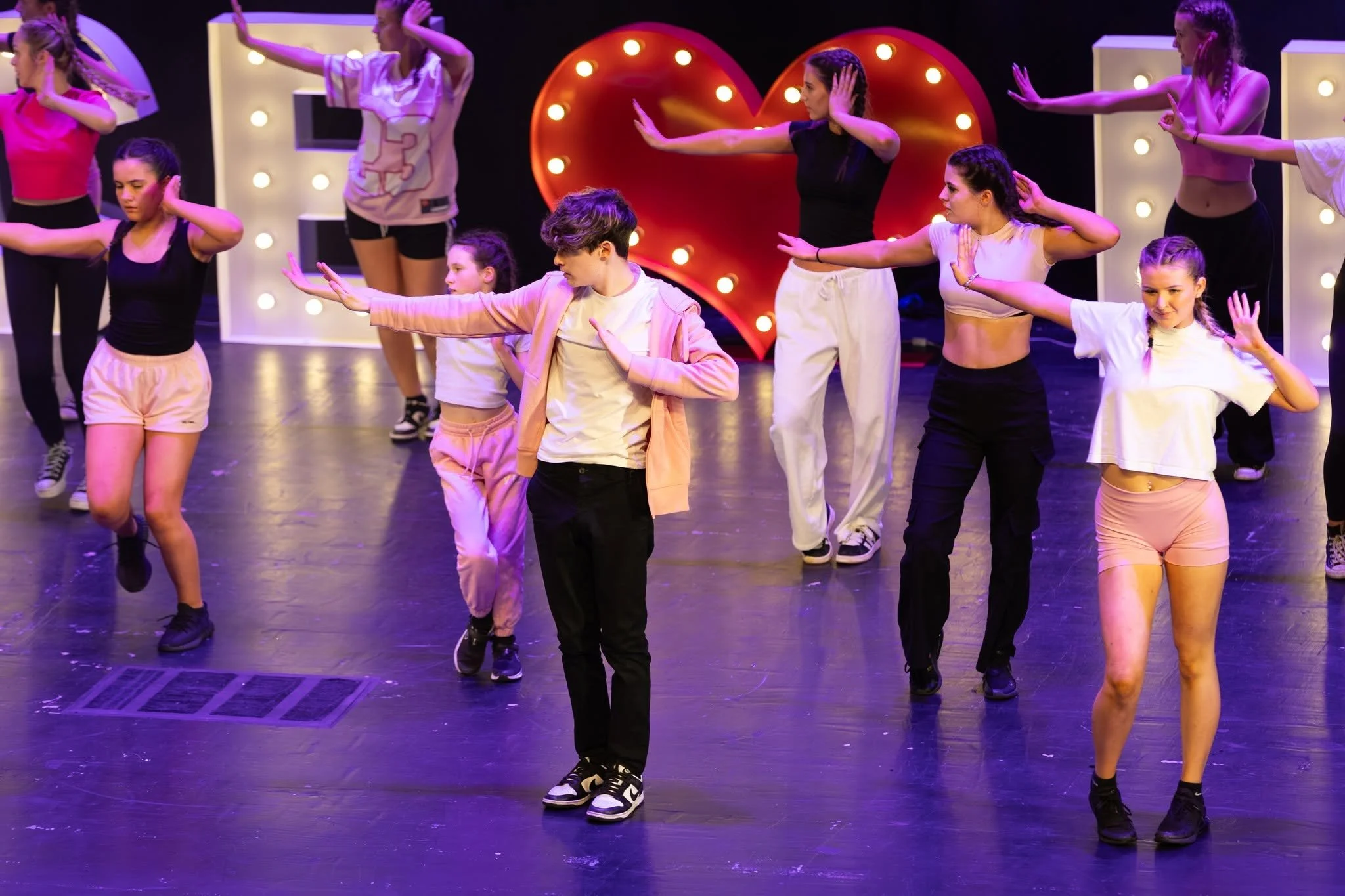 A group of young dancers performing a choreographed dance routine on a stage with decorative lights and a large illuminated heart in the background.