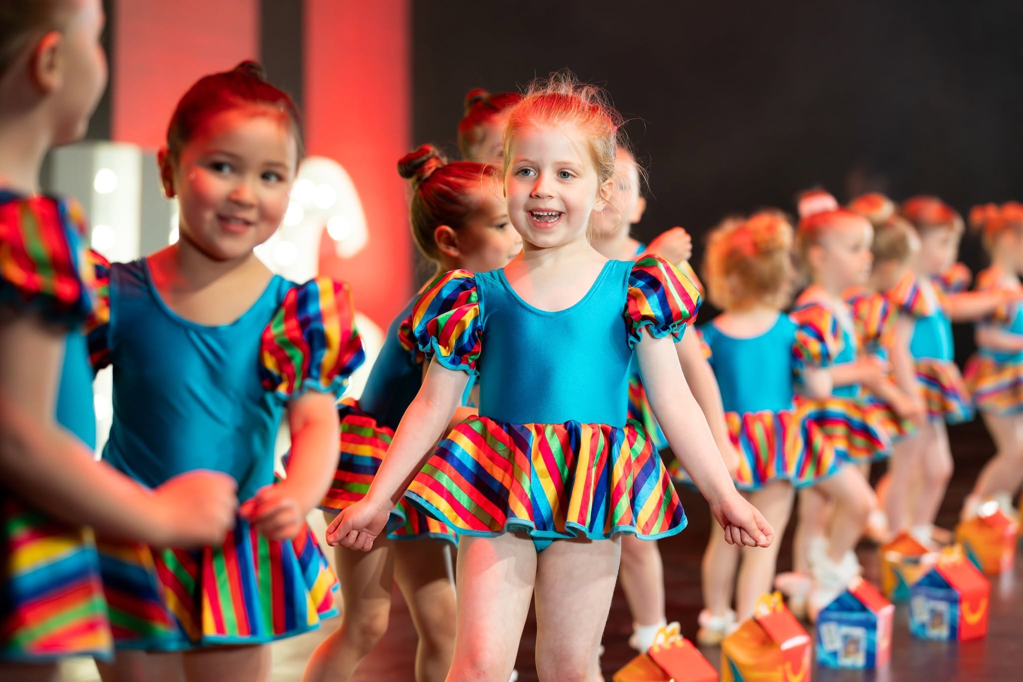 A group of young girls in colorful costumes holding hands, participating in a dance or cheerleading activity in an indoor setting.