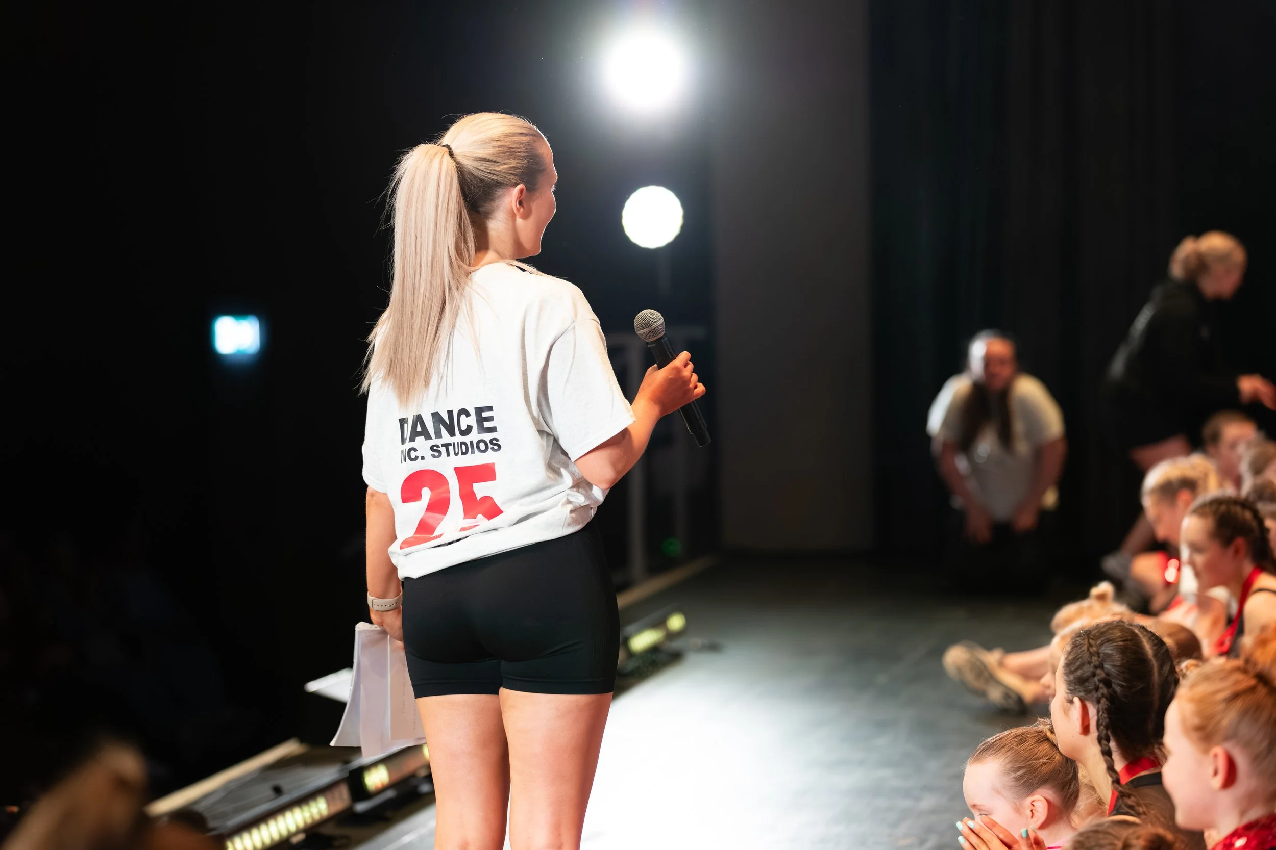 A woman with blonde hair tied in a ponytail is standing on stage holding a microphone, wearing a white t-shirt with red and black text and black shorts, with an audience of children seated in front of her.
