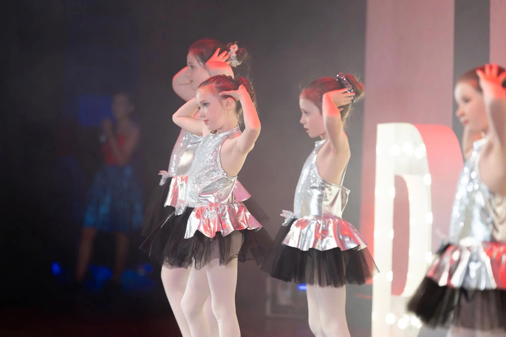 A group of young girls in silver and black tutus performing a ballet on stage, with some fixing their hair.