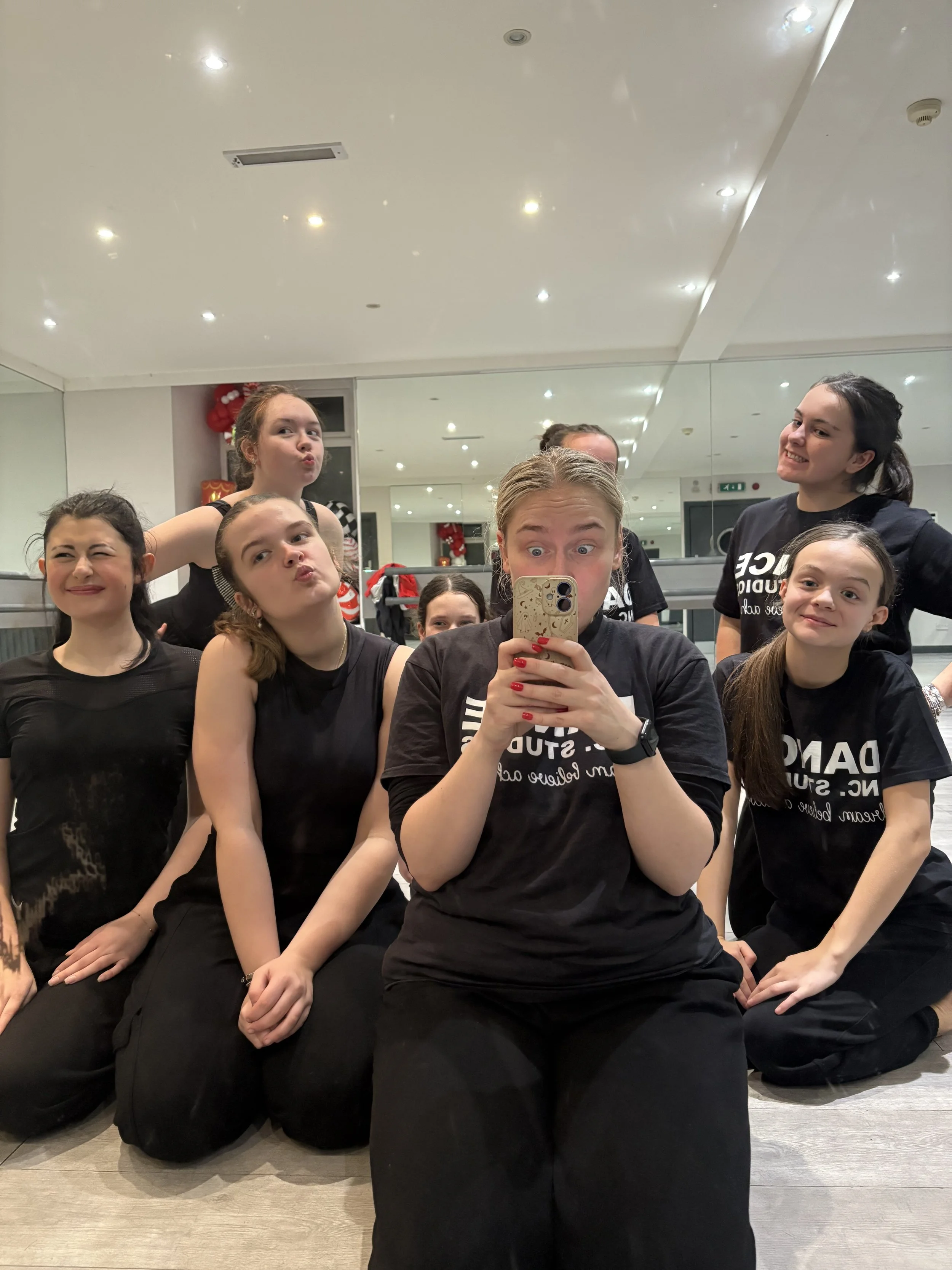 A group of young women in black shirts taking a selfie in a dance studio with mirrors, ceiling lights, and Christmas decorations in the background.