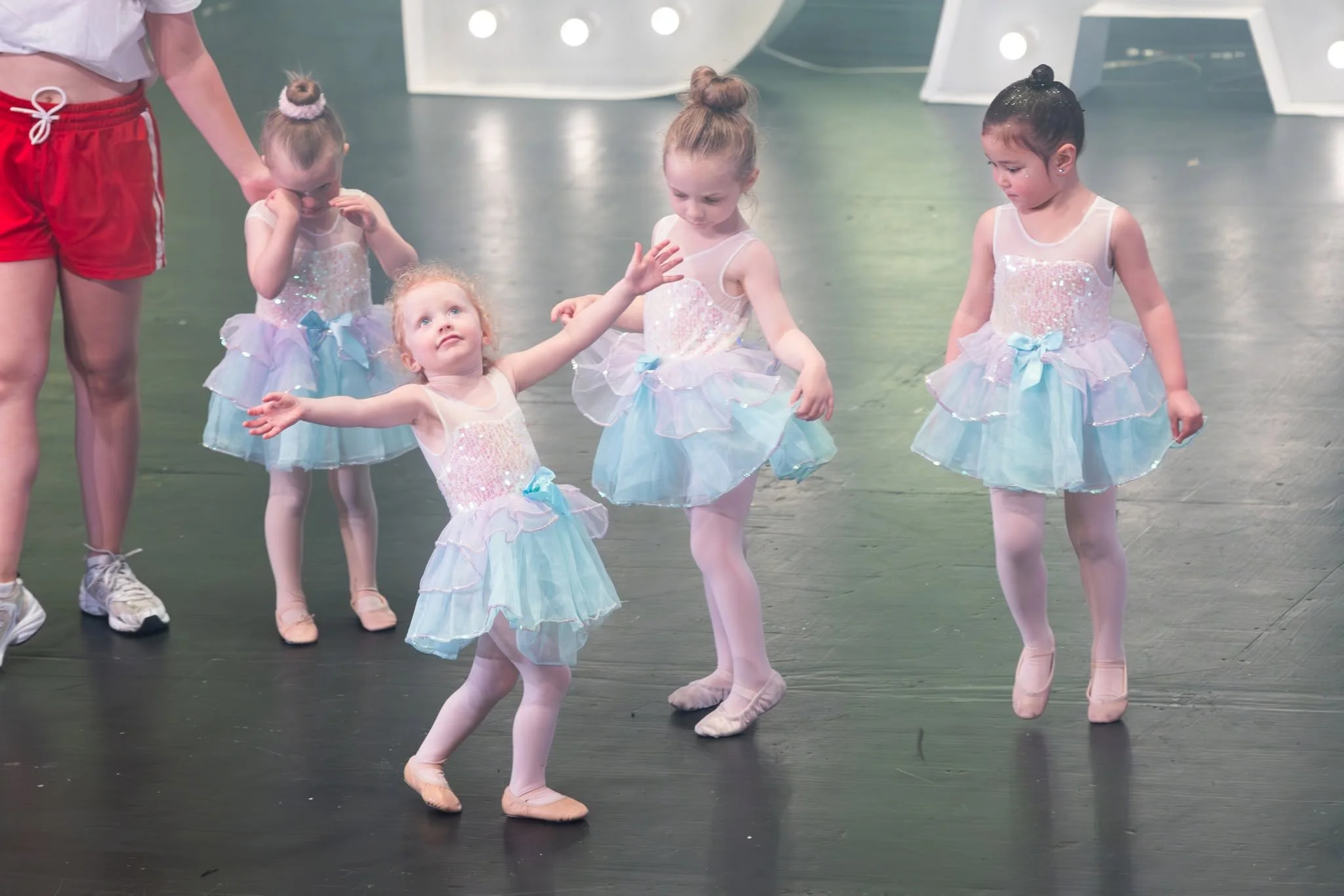 Four young girls dressed in ballet costumes practicing dance moves on a stage, with a coach or instructor nearby.