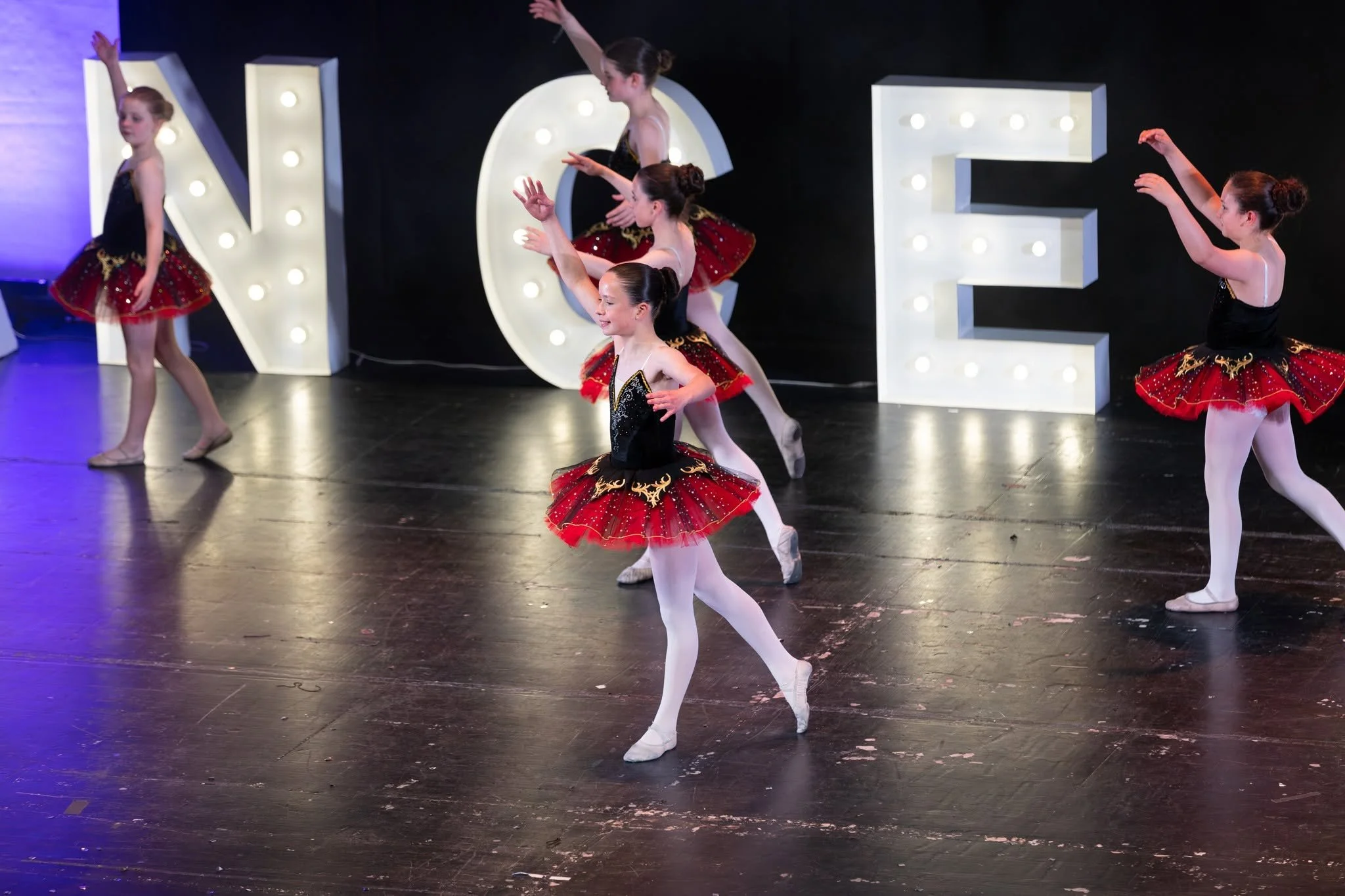 Young girls in black dance costumes with pink ruffles practicing on stage under purple lighting.