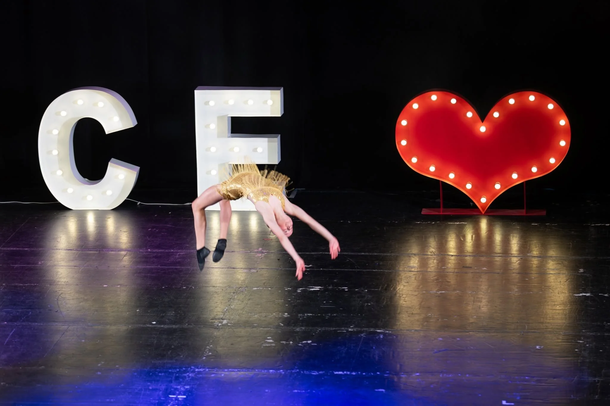 A group of dancers performing on stage, with one dancer in the foreground stretching with arms outstretched and head tilted back, wearing a black sequin top, and a black tutu with a pink bow.