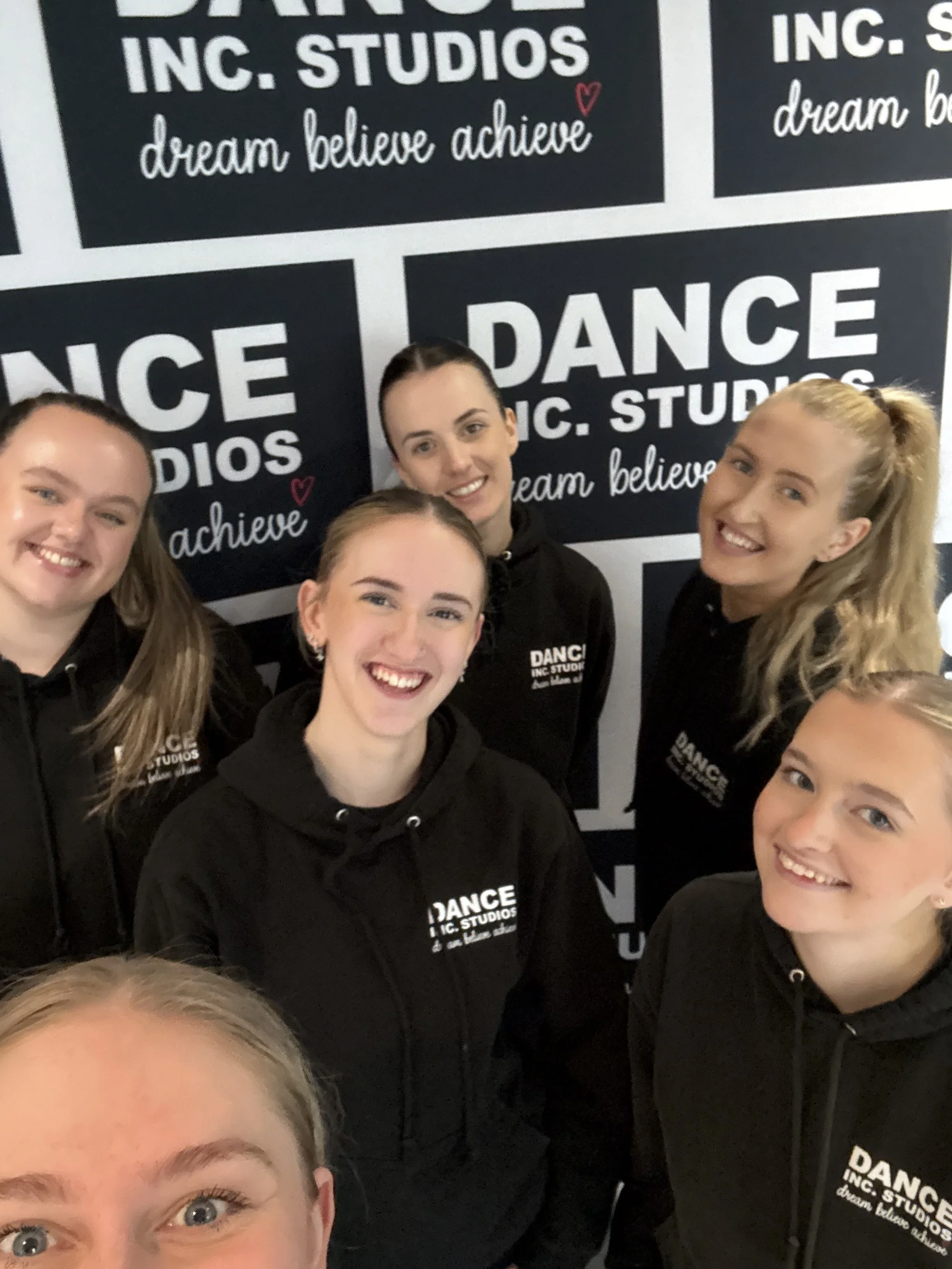 Group of smiling young women in black sweatshirts posing for a photo indoors, standing in front of a black and white wall with the words 'DANCE INC. STUDIOS' and the phrases 'dream believe achieve' and 'dance' visible.