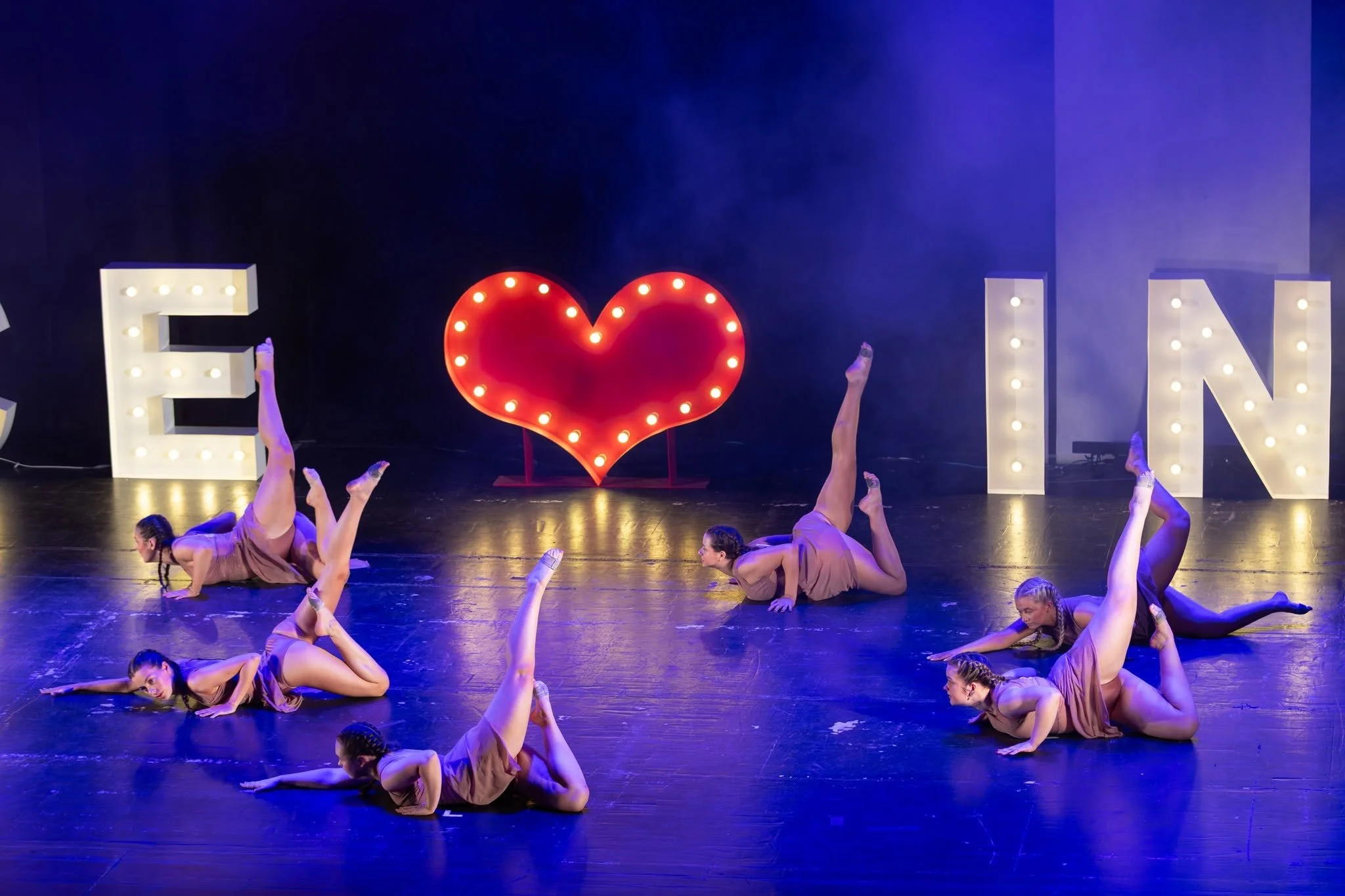 Four young girls dressed in ballet costumes practicing dance moves on a stage, with a coach or instructor nearby.