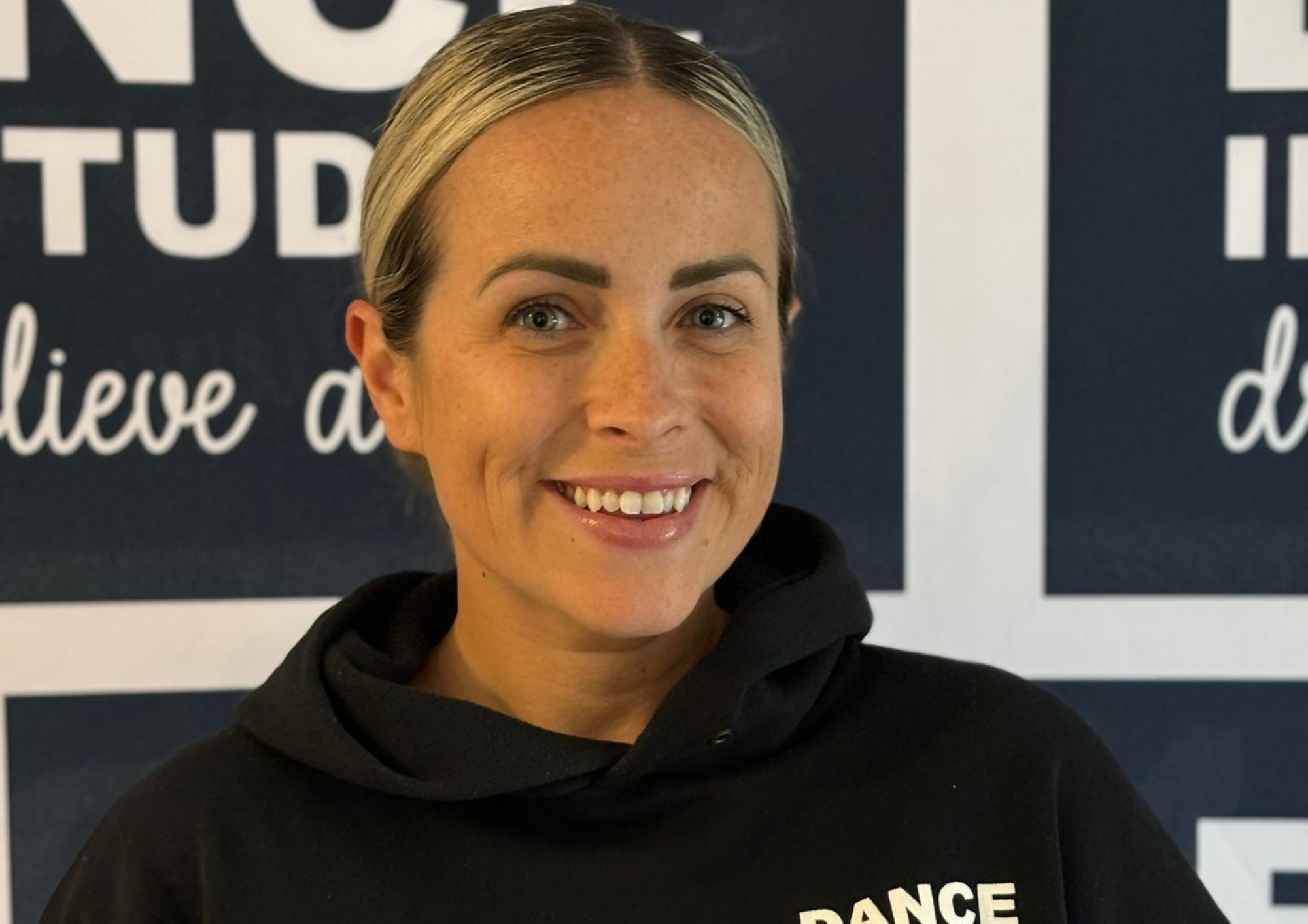 A smiling woman with short light hair and light skin taking a selfie in front of a dark background with white text.