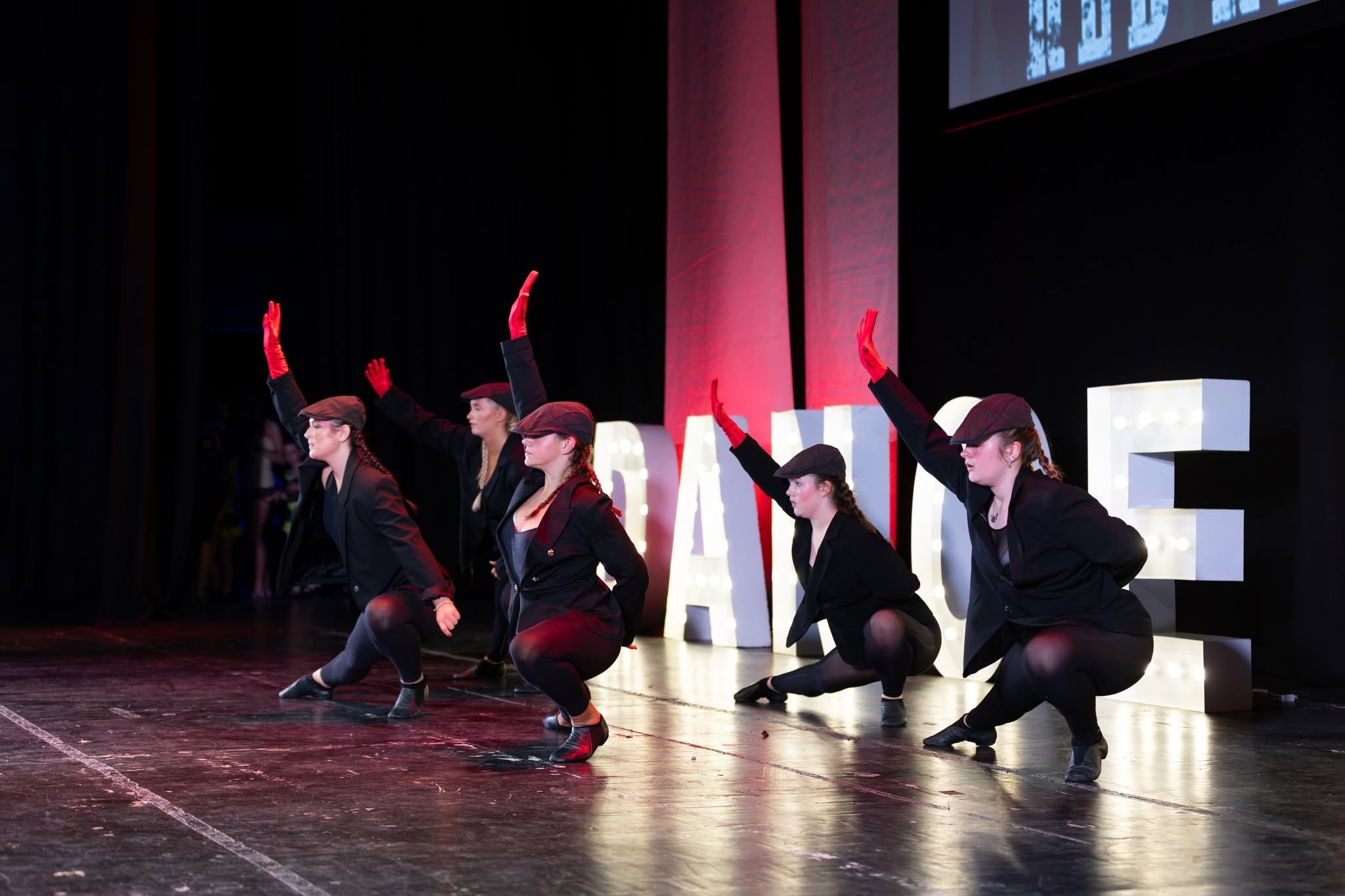 Children performing breakdance on stage, wearing yellow safety vests and white pants, with a dark background and a group of young women in casual clothes standing in the background.