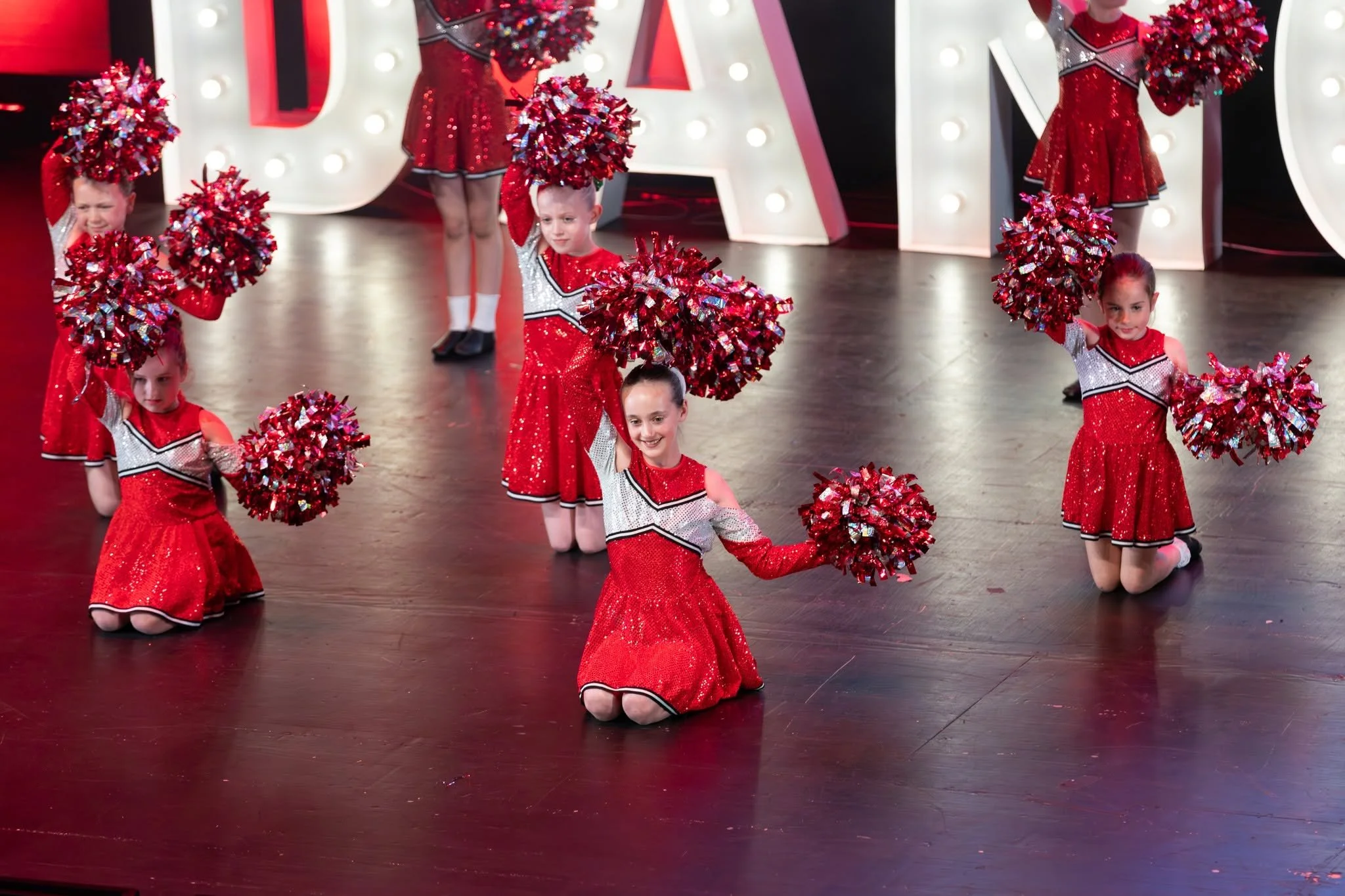 Young girls performing a cheerleading routine on stage dressed in red sparkly cheerleader outfits with shiny pom-poms.