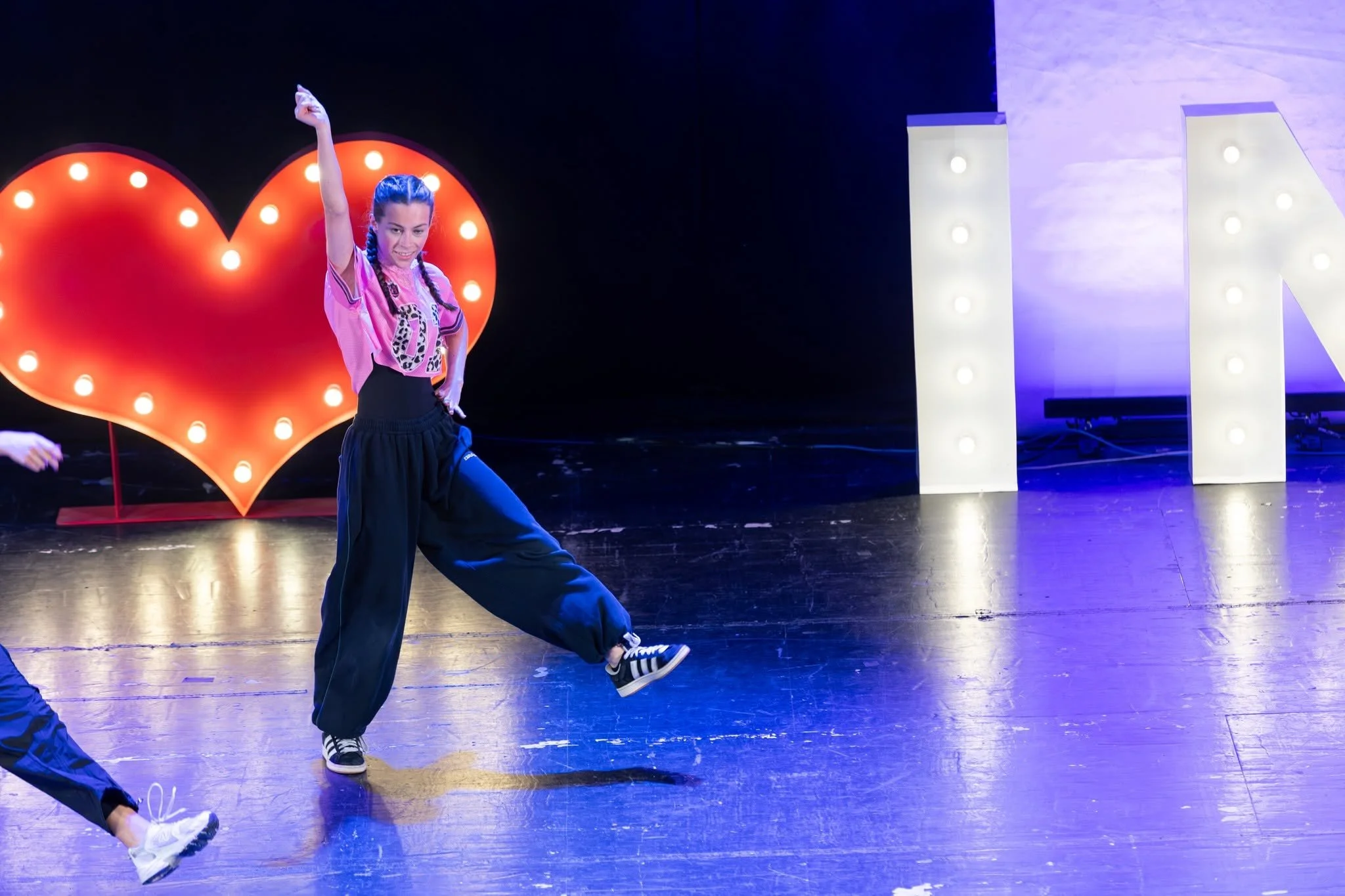 Group of female dancers performing a choreographed routine on stage with large illuminated letters in the background.
