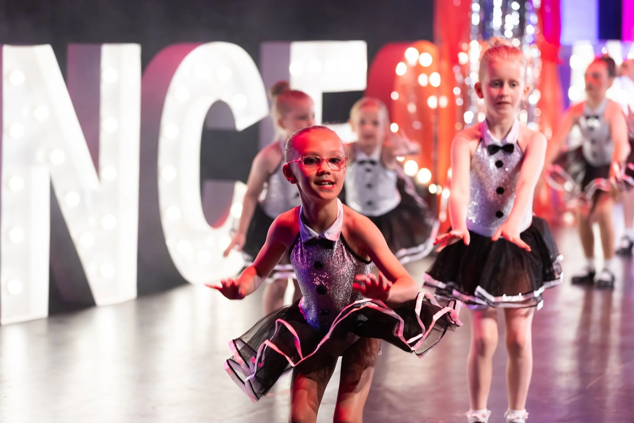Group of young girls in dance costumes performing a dance routine on stage with illuminated sign in background