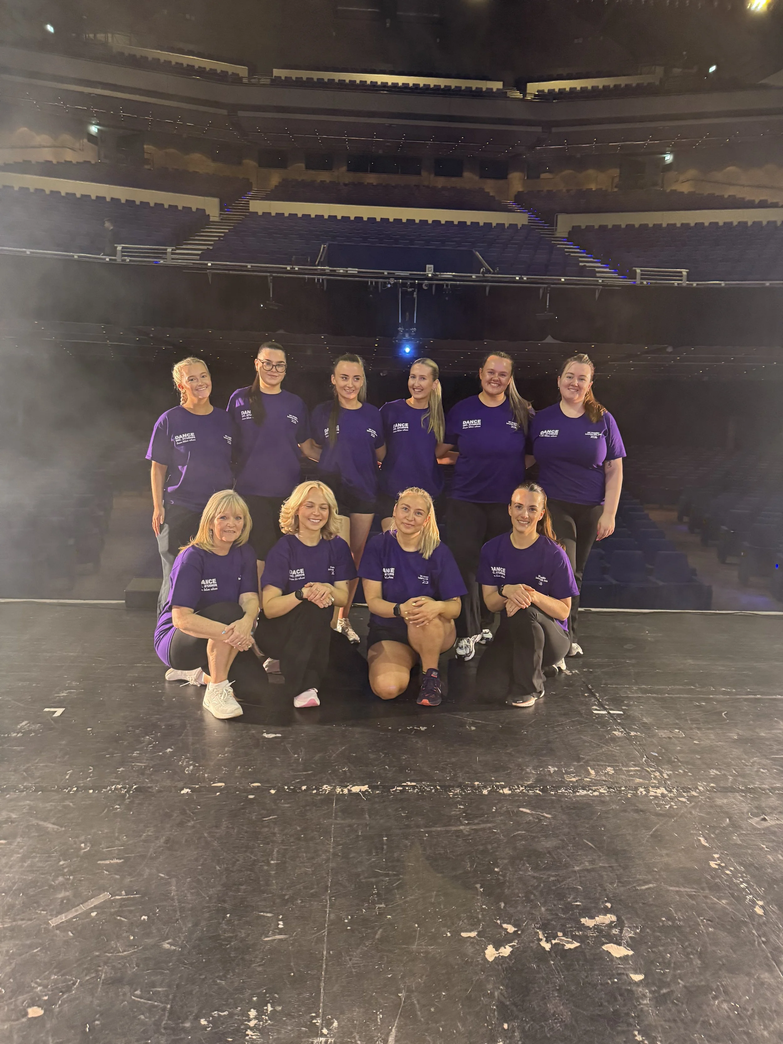 A group of women in purple dance team shirts posing on a stage in an empty theater or auditorium with rows of seats and balconies in the background.