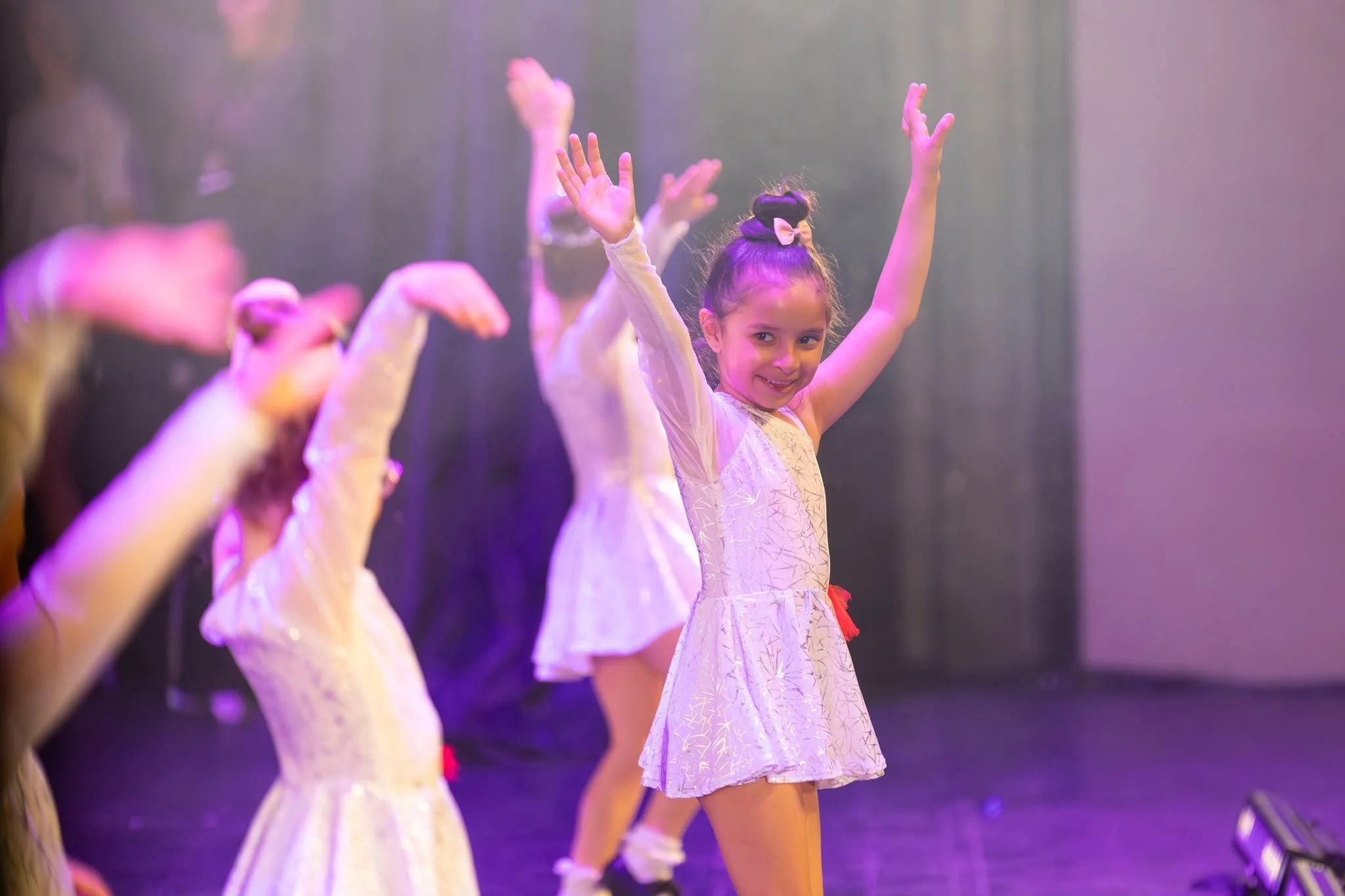 Young girl in a light-colored dress with a pink bow in her hair dancing on stage with others, raising her arms.