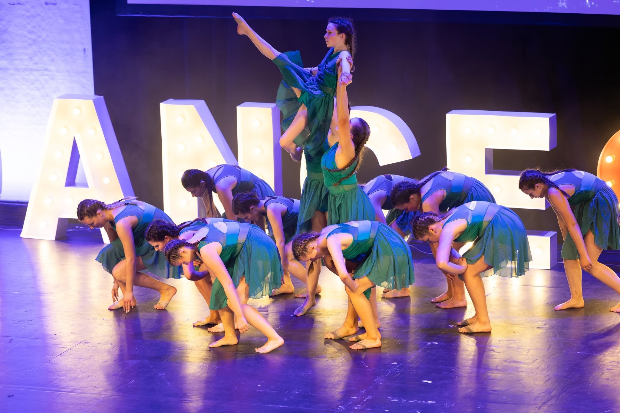 Group of female dancers performing a choreographed routine on stage with large illuminated letters in the background.