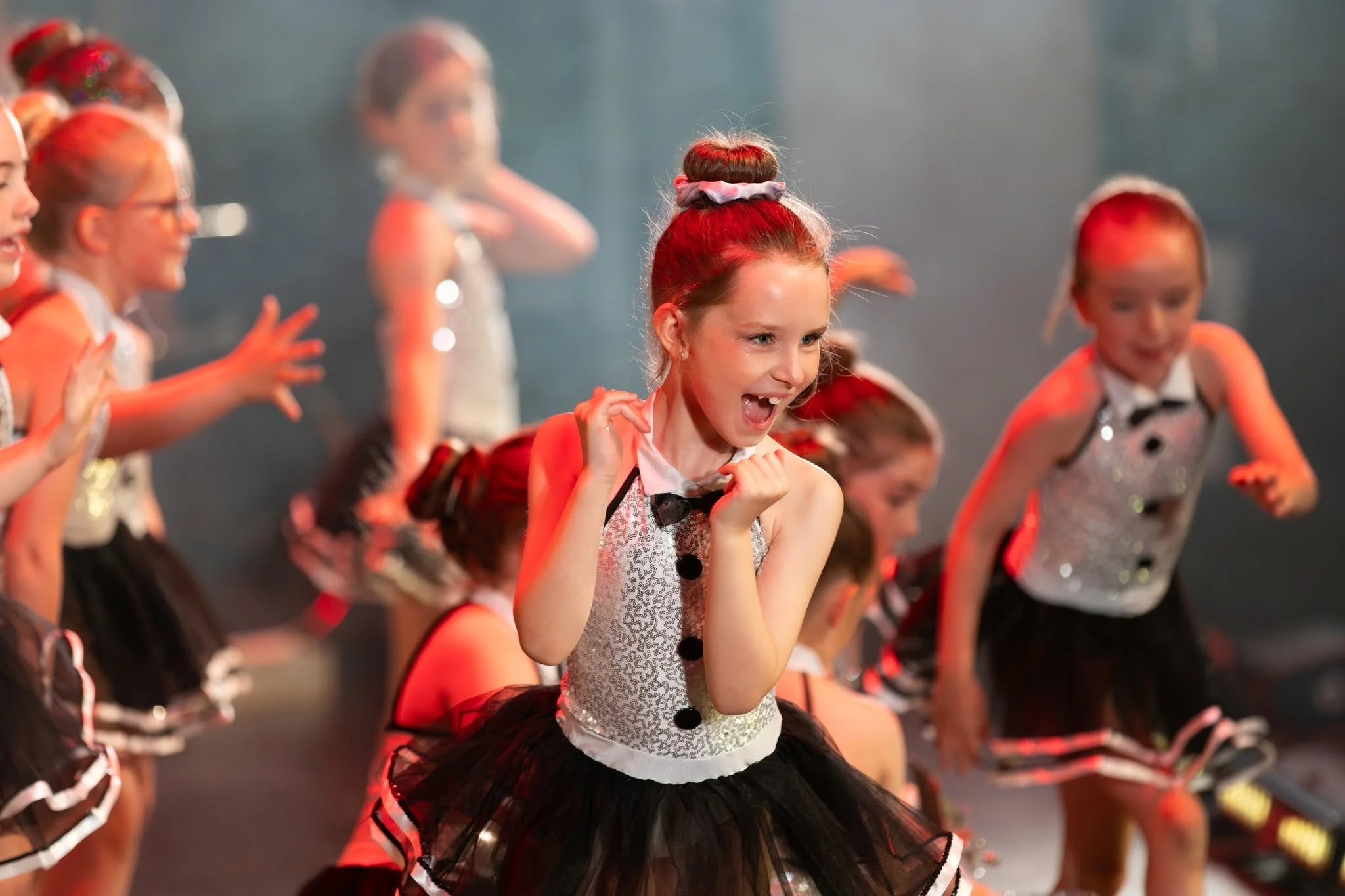 Young girl with red hair in a bun, wearing a sparkly white top with black buttons and a black tulle skirt, smiling and dancing with other children in colorful costumes on stage.