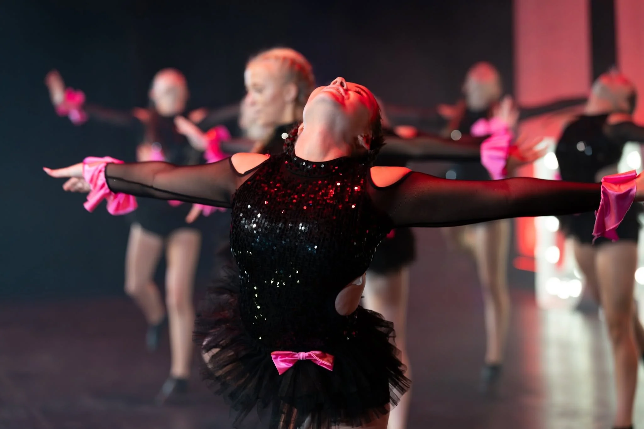 A group of dancers performing on stage, with one dancer in the foreground stretching with arms outstretched and head tilted back, wearing a black sequin top, and a black tutu with a pink bow.